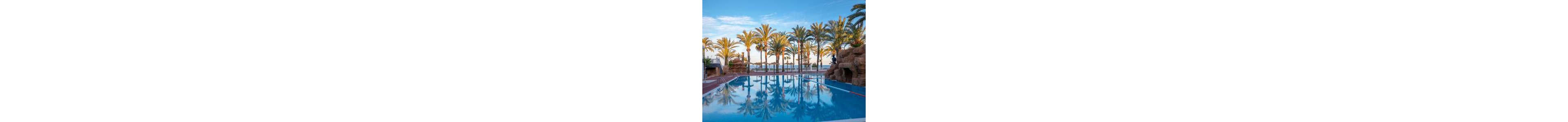 A clear blue swimming pool reflects tall palm trees under a soft morning or evening sky. The poolside area is lined with red tiles and bordered by artificial rock formations and a white balustrade. Beyond the pool, the sea and beach umbrellas suggest a coastal resort setting. The calm water and warm light create a tranquil vacation atmosphere.