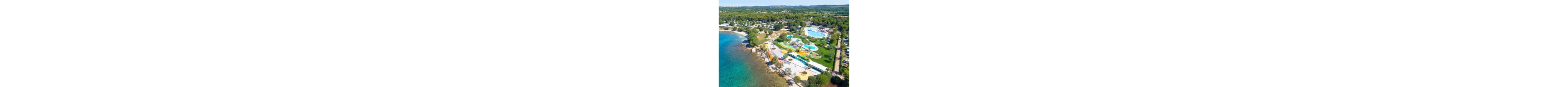 A large coastal resort with a colorful water park is captured from an aerial perspective. Multiple pools, winding slides, and play structures are arranged near the shoreline beside clear blue water. Campsites and buildings extend into the surrounding green landscape, indicating a spacious holiday area. The bright colors and sunny weather create a lively, family-friendly vacation atmosphere.
