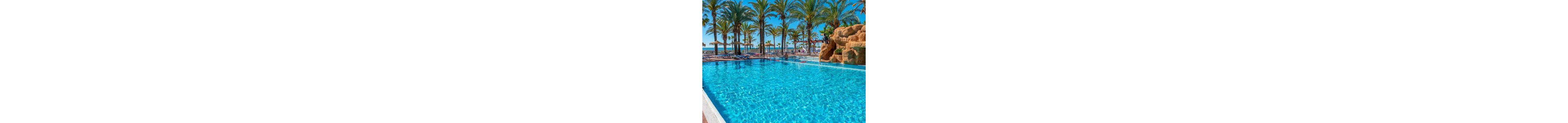 A bright blue swimming pool sparkles under clear sunlight at a tropical resort. Tall palm trees line the background, framing lounge chairs and sun umbrellas along the poolside. A rock feature with a small waterfall structure adds interest on the right side. The scene suggests a relaxing vacation atmosphere by the water on a sunny day.