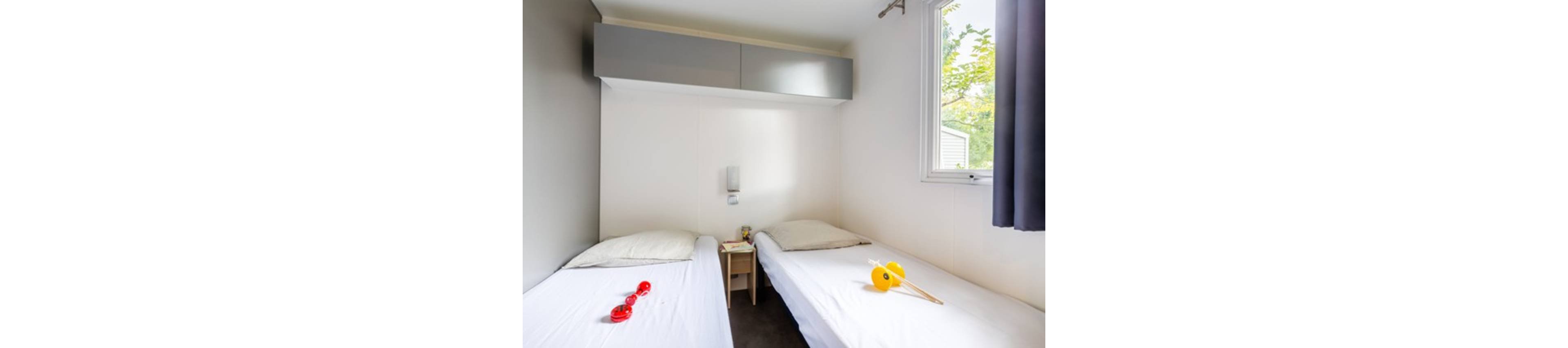 A small bedroom features two single beds separated by a narrow wooden nightstand. The room has white walls, a large window with a curtain, and overhead storage cabinets above one bed. Simple pillows rest on each bed, while colorful toys or objects lie on the blankets. Natural light from the window brightens the clean, minimalist space and offers a view of greenery outside.