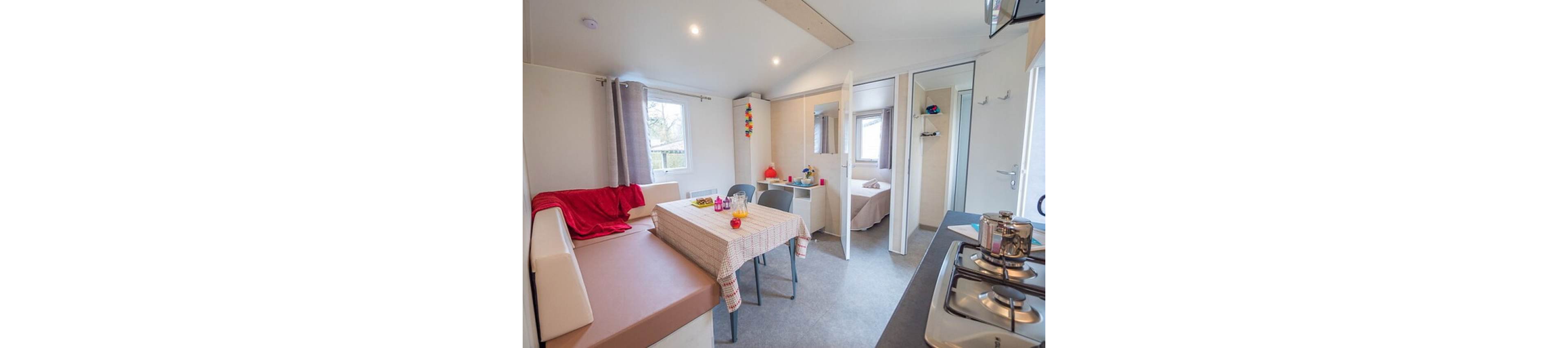 Cozy mobile home interior featuring a small dining area with a checkered tablecloth and plastic chairs. A built-in bench with a red blanket lines one wall, while a compact kitchen with a gas stove and kettle sits opposite. The space is brightly lit with recessed ceiling lights and natural light from multiple windows with curtains. An open doorway reveals a small bedroom, emphasizing the compact yet functional vacation accommodation.