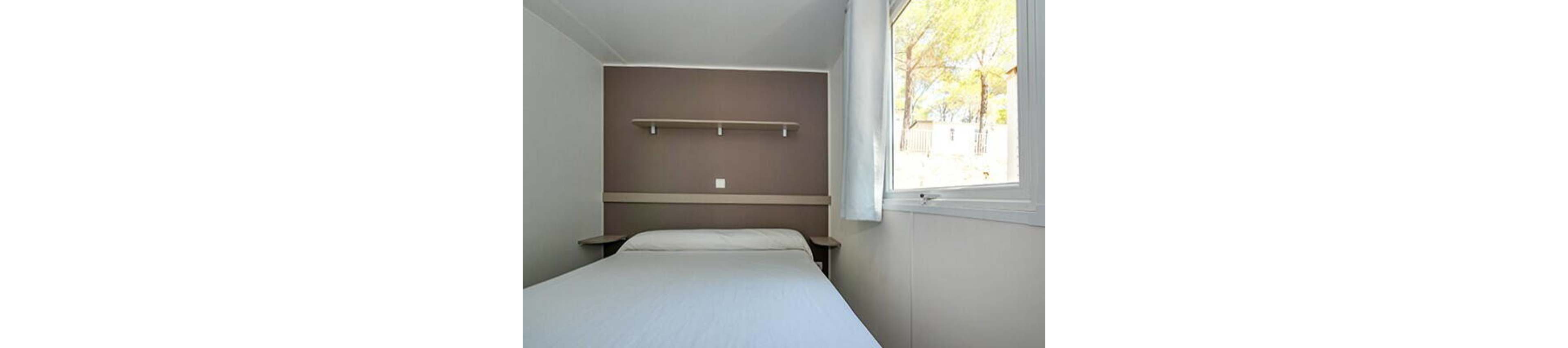A compact bedroom features a single bed neatly made with white bedding. The room has a brown accent wall with a simple floating shelf and a central power outlet. Natural light enters through a large window with white curtains, revealing trees and an outdoor area. The overall design is clean, minimal, and functional, suitable for a small apartment or vacation cabin.