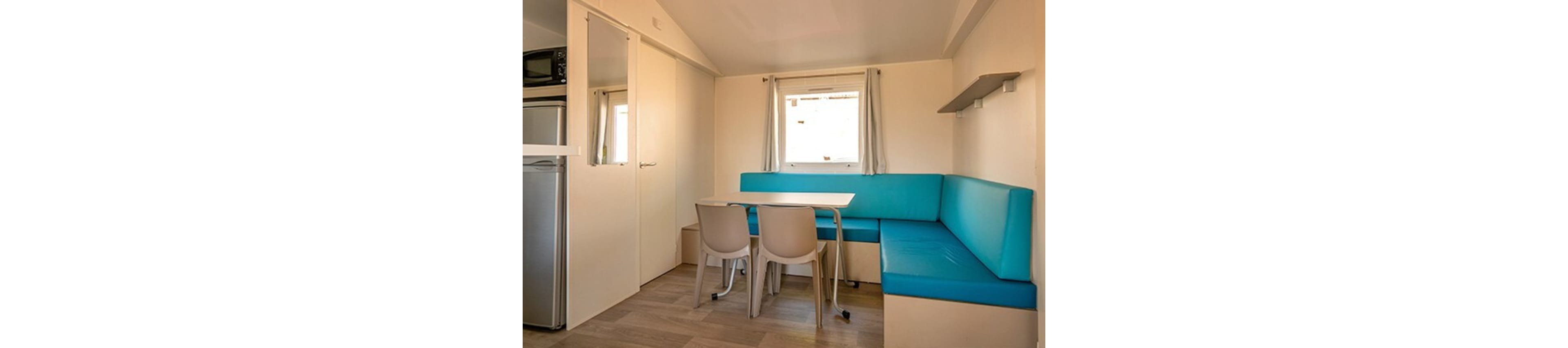 A compact dining and seating area is shown inside a small modern apartment or mobile home. A turquoise upholstered corner bench wraps around a simple white dining table with beige plastic chairs. Light wood flooring and cream-colored walls create a bright, minimalist interior. A small window with light curtains and built-in shelves add functionality to the cozy space.