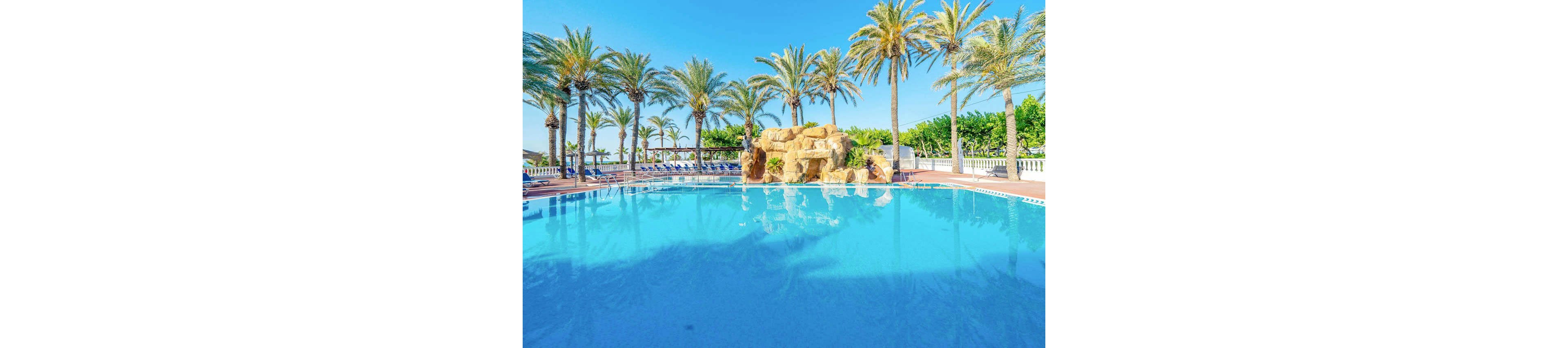 A large, clear blue swimming pool reflects the bright sky at a sunny resort. Tall palm trees line the poolside, creating a tropical atmosphere. A rocky waterfall feature stands at the far end of the pool, with lounge chairs and umbrellas visible in the background. The scene suggests a relaxing vacation setting on a warm, cloudless day.