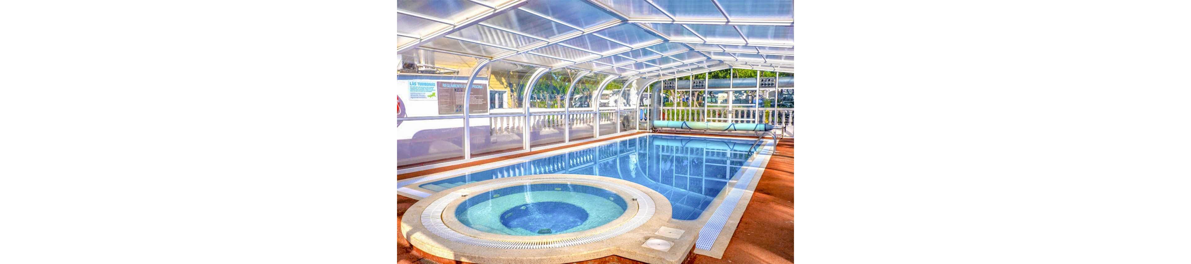 A bright indoor swimming pool is enclosed by a transparent arched structure. In the foreground, a circular hot tub with bubbling water sits beside the main rectangular pool. Sunlight filters through the clear roof, reflecting off the calm blue water. White railings and outdoor greenery are visible through the glass walls, creating a relaxing resort-like atmosphere.