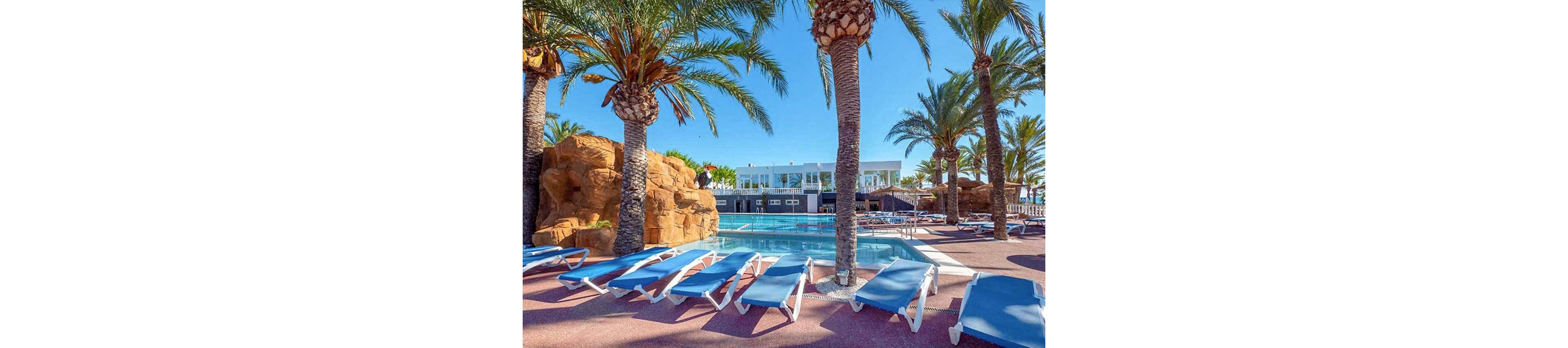 An outdoor swimming pool is surrounded by rows of blue sun loungers under tall palm trees. A white resort building stands in the background, overlooking the pool area. Rocky formations and clear blue water create a tropical vacation atmosphere. The bright daylight and clear sky emphasize a warm, relaxing holiday setting.