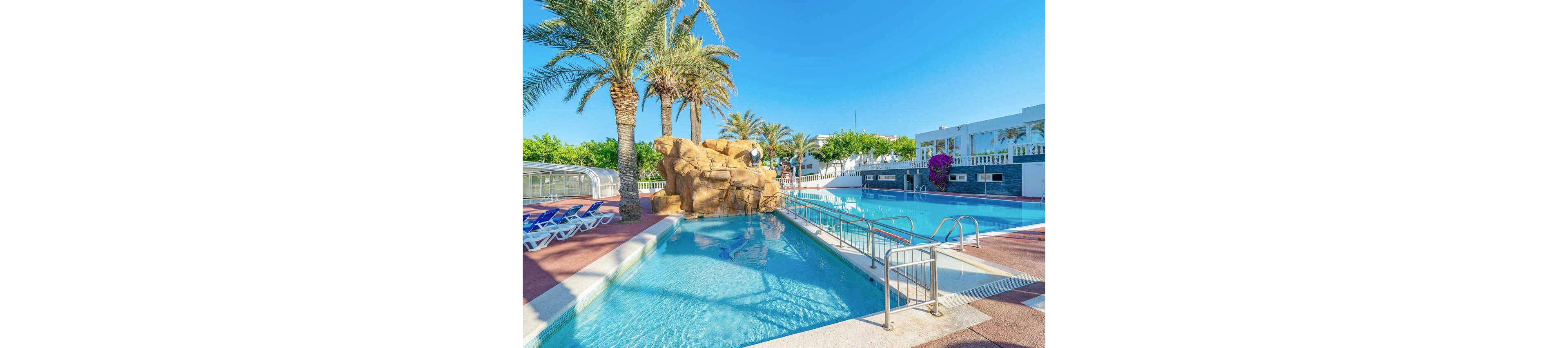 An outdoor resort swimming pool is surrounded by tall palm trees under a clear blue sky. The pool area features a rock formation in the water and metal ladders for easy access. White lounge chairs line the deck, and white buildings with balconies and railings frame the background. Bright sunlight and vivid colors create a warm, inviting vacation atmosphere.