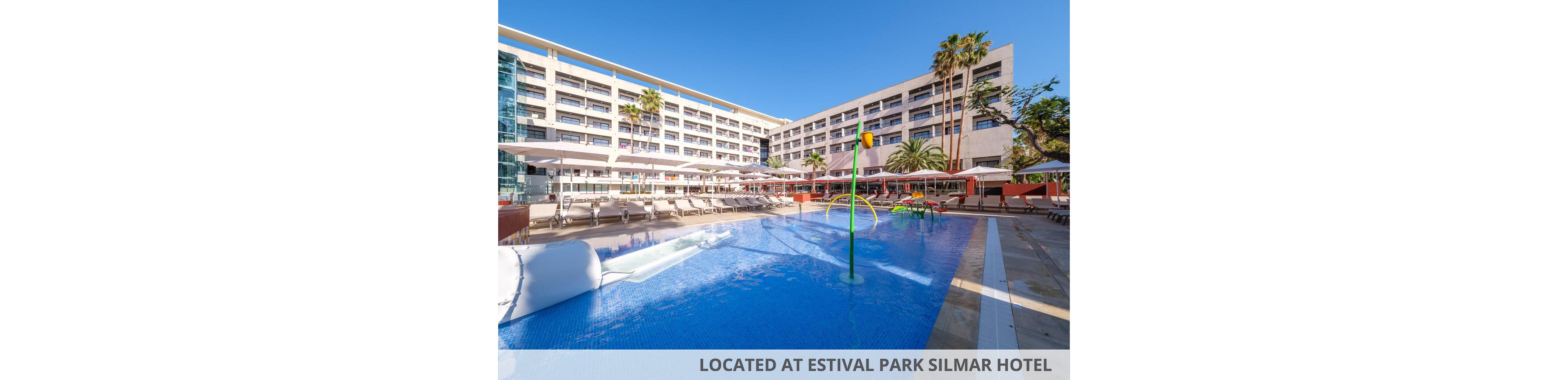 A large outdoor swimming pool is shown in front of the Estival Park Silmar hotel complex. The multi-story hotel building features numerous balconies and is surrounded by palm trees and sun loungers. Colorful water play structures stand in the shallow pool area under a clear blue sky. A caption at the bottom reads "LOCATED AT ESTIVAL PARK SILMAR HOTEL."