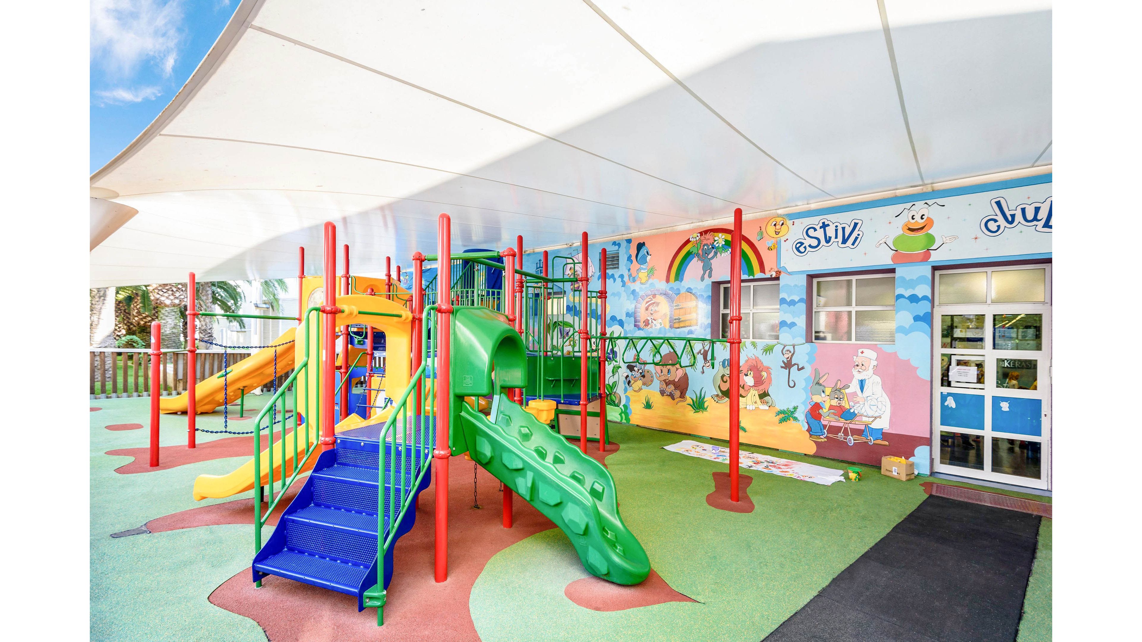 A bright outdoor playground features a green and yellow slide structure with red support poles. The play area is covered by a large white canopy and has soft, multicolored safety flooring. A mural with cartoon animals, rainbows, and playful scenes decorates the wall beside the equipment. The space appears clean, safe, and inviting for young children at a daycare or preschool.