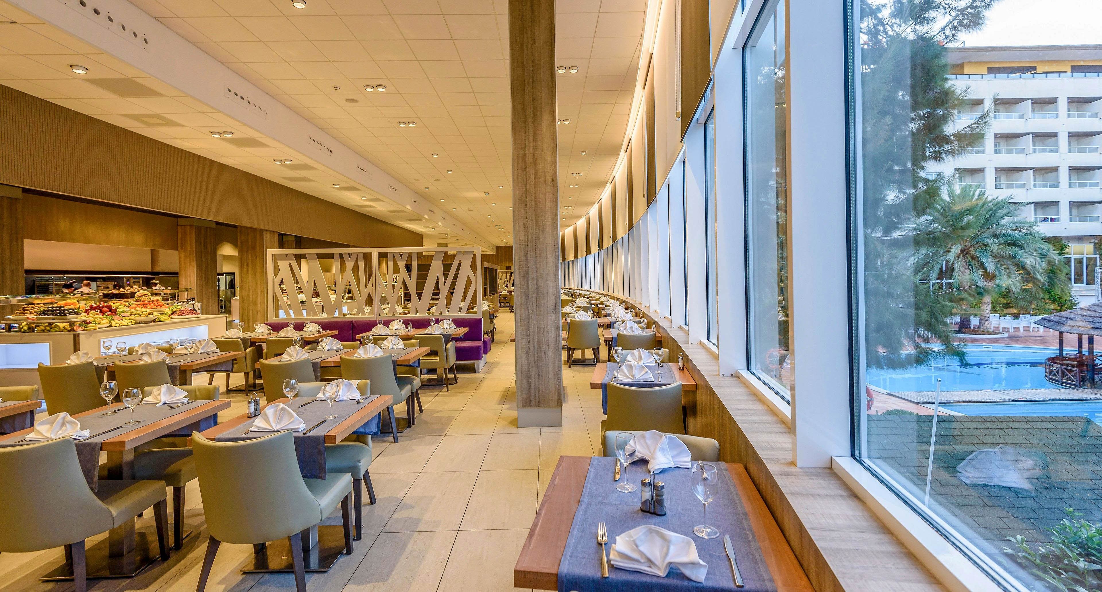 A contemporary hotel restaurant features neatly arranged tables with folded napkins and glassware. Large floor-to-ceiling windows line one side, offering a clear view of an outdoor swimming pool and surrounding palm trees. Soft neutral tones, wooden accents, and upholstered chairs create a warm, inviting atmosphere. The space appears bright and spacious, suitable for casual or formal dining.