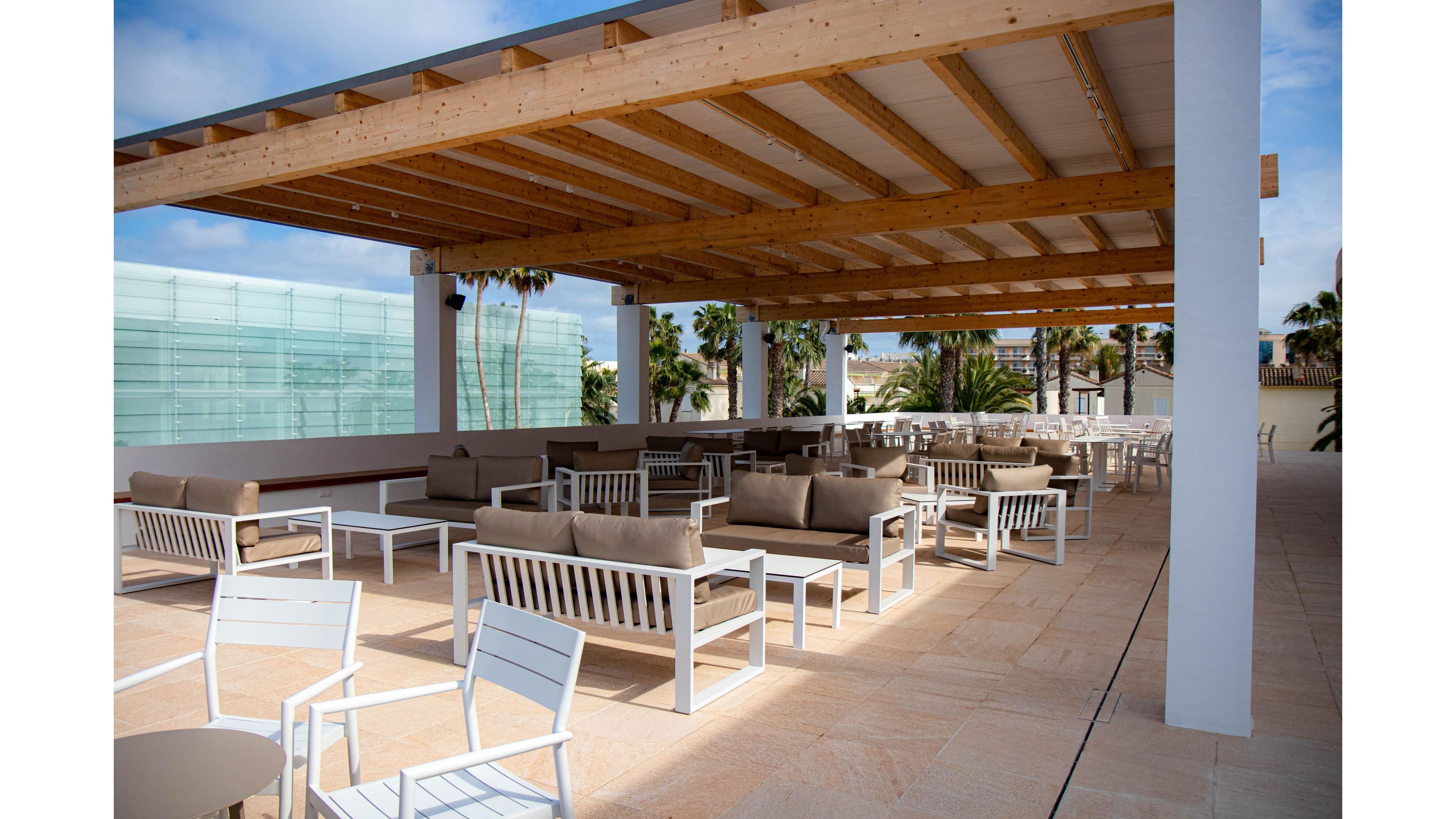 An open-air terrace features modern lounge furniture arranged beneath a large wooden pergola. Neutral-toned sofas and white chairs surround low tables on a light stone floor. Palm trees and contemporary glass architecture are visible in the background, suggesting a coastal or resort setting. The space appears calm and inviting, suitable for relaxation, social gatherings, or outdoor dining.