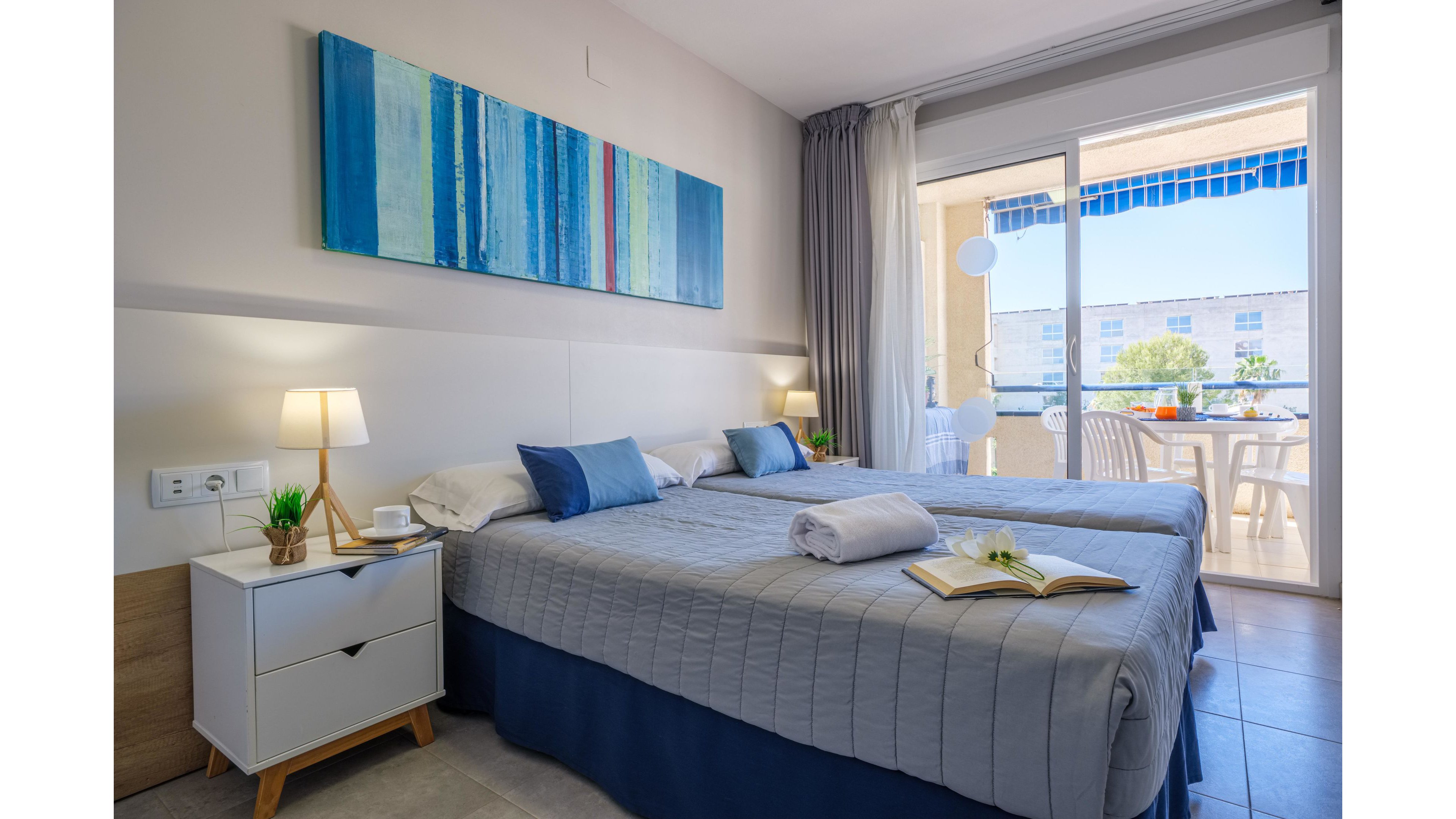 A modern hotel bedroom features two neatly made twin beds with gray quilts and blue accent pillows. A white nightstand with a lamp, plant, and cup sits beside the bed, while a large abstract blue painting decorates the wall above. Sunlight streams in through sliding glass doors that open onto a balcony with white outdoor furniture and a view of nearby buildings and trees. The overall scene feels clean, inviting, and ideal for a relaxing vacation stay.