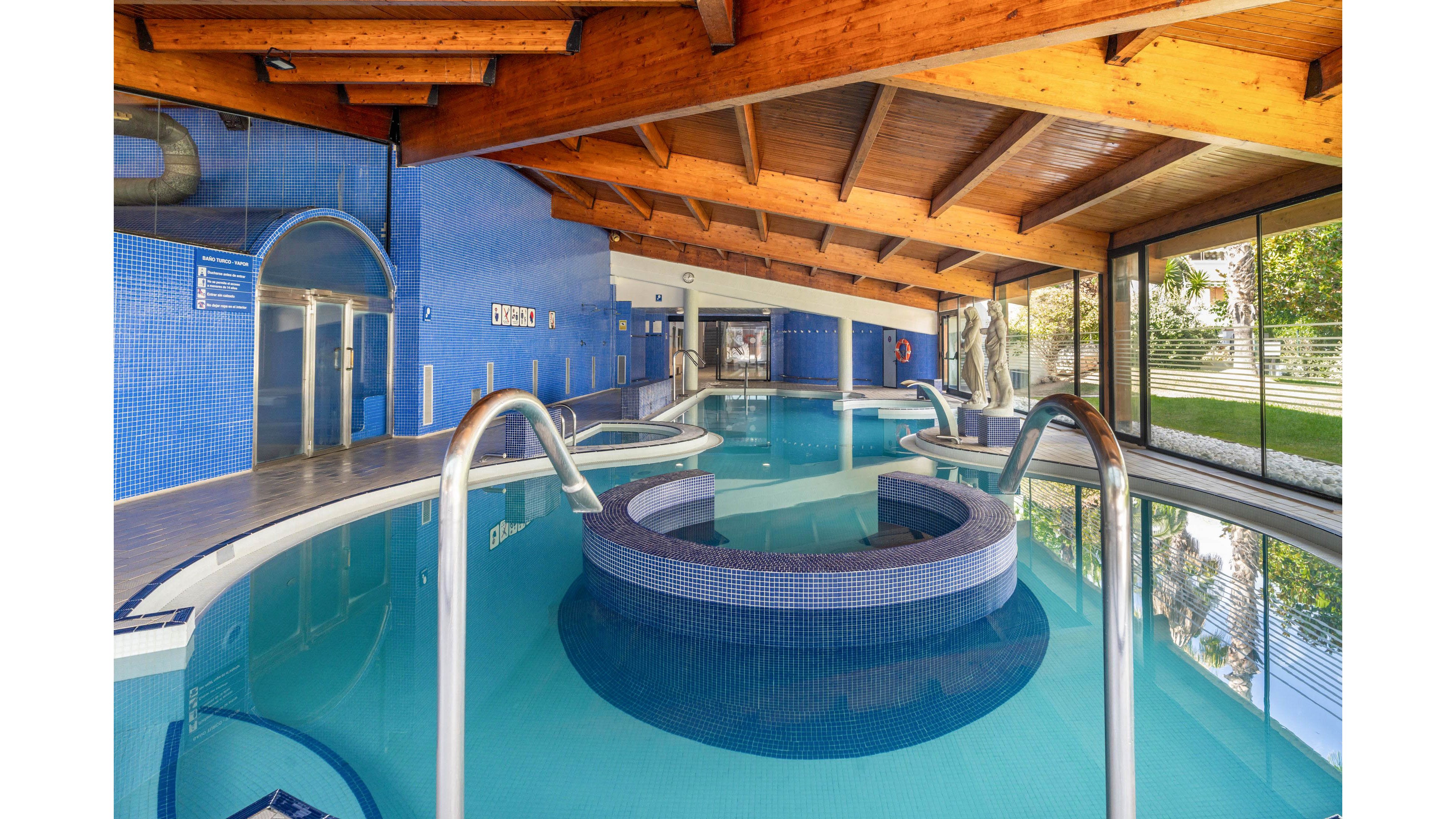 A contemporary indoor spa pool area features curved pools and circular whirlpool sections lined with blue mosaic tiles. Stainless steel handrails and steps lead into the water beneath a warm wooden beam ceiling. Large windows and glass doors open to a green outdoor garden, bringing natural light into the serene space. The scene suggests a relaxing wellness or resort environment designed for comfort and leisure.