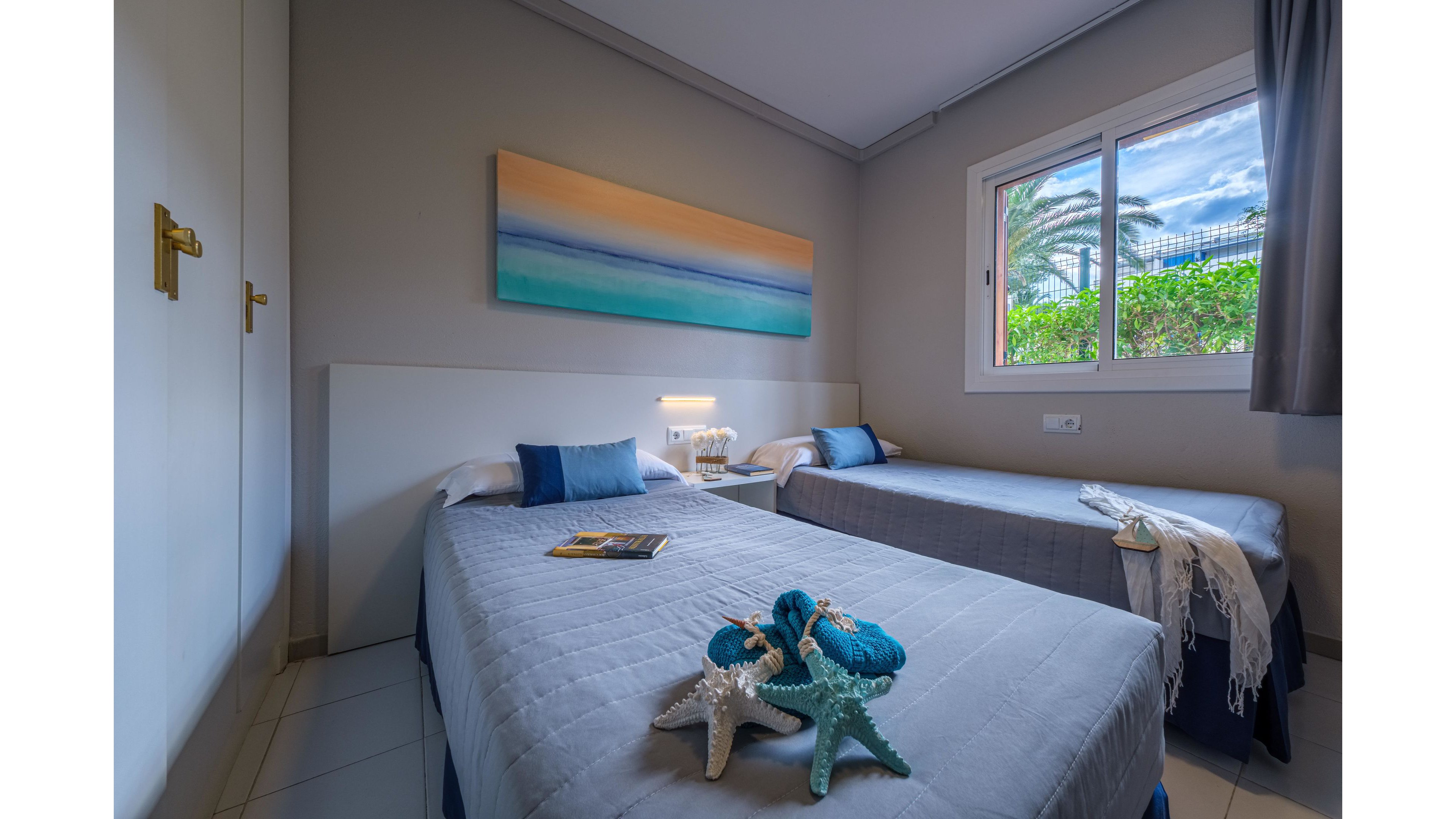 A small bedroom features two neatly made twin beds with gray bedspreads and white headboards. Nautical decorations, including starfish and blue rope, are arranged on the nearest bed, adding a coastal touch. A large abstract seascape painting hangs above the beds, complementing the blue and beige color palette. Natural light enters through the window, revealing greenery and palm trees outside, while a bedside table and lamp provide additional comfort.