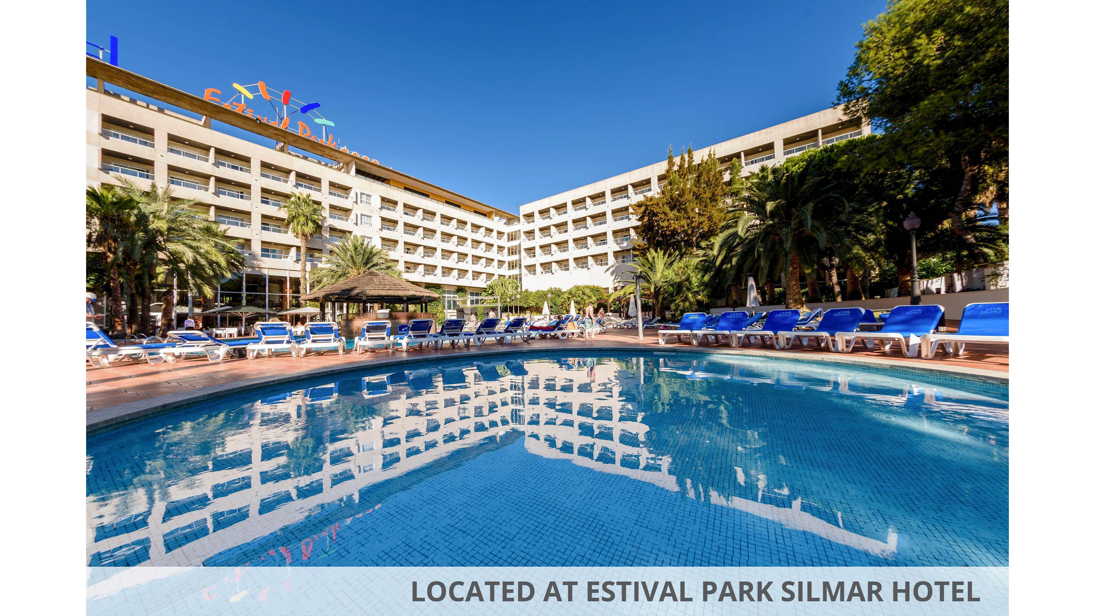 Large resort hotel with multiple balconies overlooks a clear blue outdoor swimming pool. Blue sun loungers and umbrellas surround the poolside terrace under bright sunny skies. Palm trees and lush greenery frame the leisure area, creating a relaxed vacation atmosphere. Text at the bottom reads "LOCATED AT ESTIVAL PARK SILMAR HOTEL."