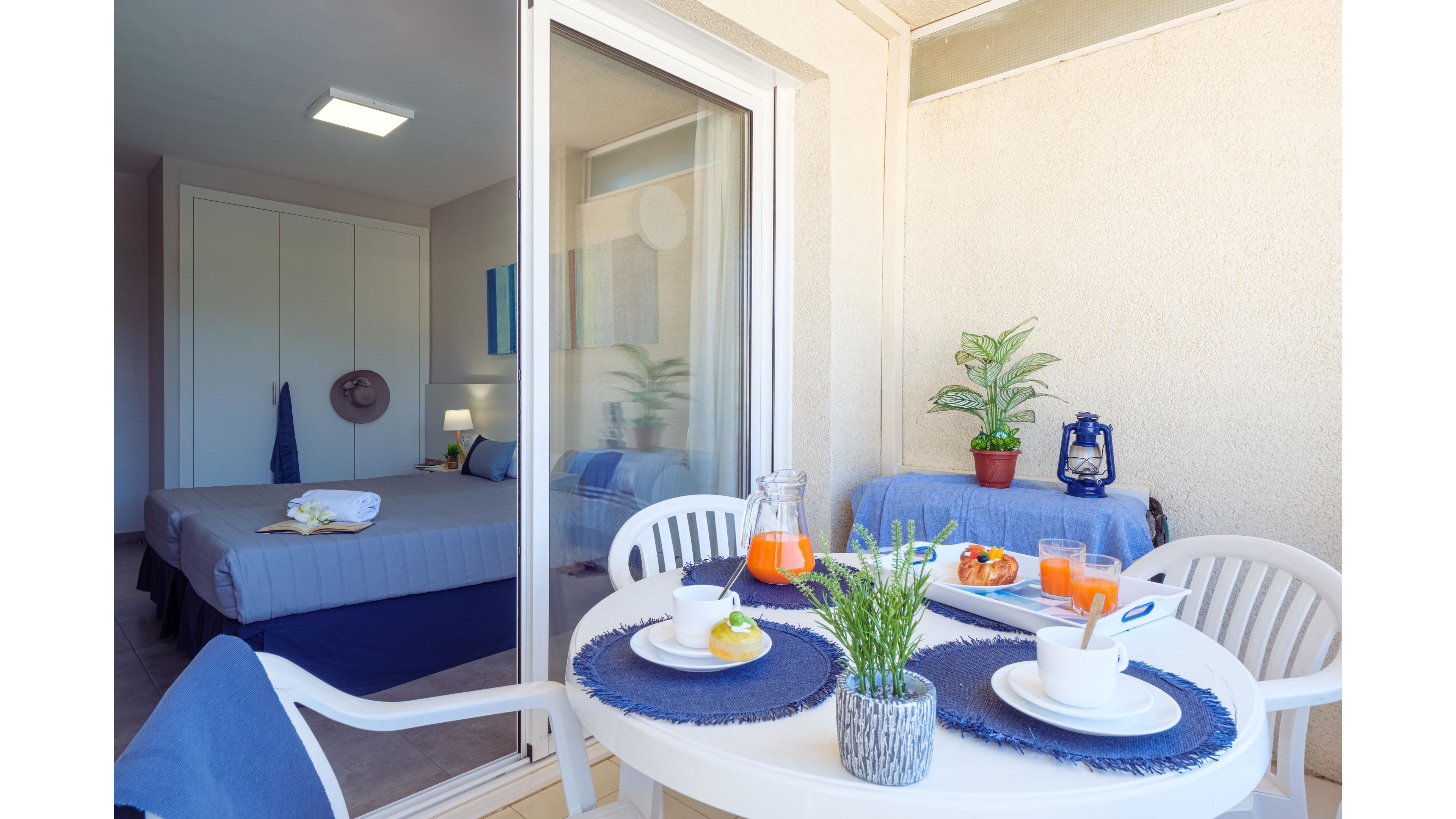 A bright balcony dining area is set with a round white table and blue placemats. Breakfast items including juice, pastries, and coffee are arranged neatly, creating an inviting scene. Through the sliding glass door, a tidy bedroom with a made bed and wardrobe is visible. Potted plants and soft neutral walls add a relaxed, holiday atmosphere to the space.