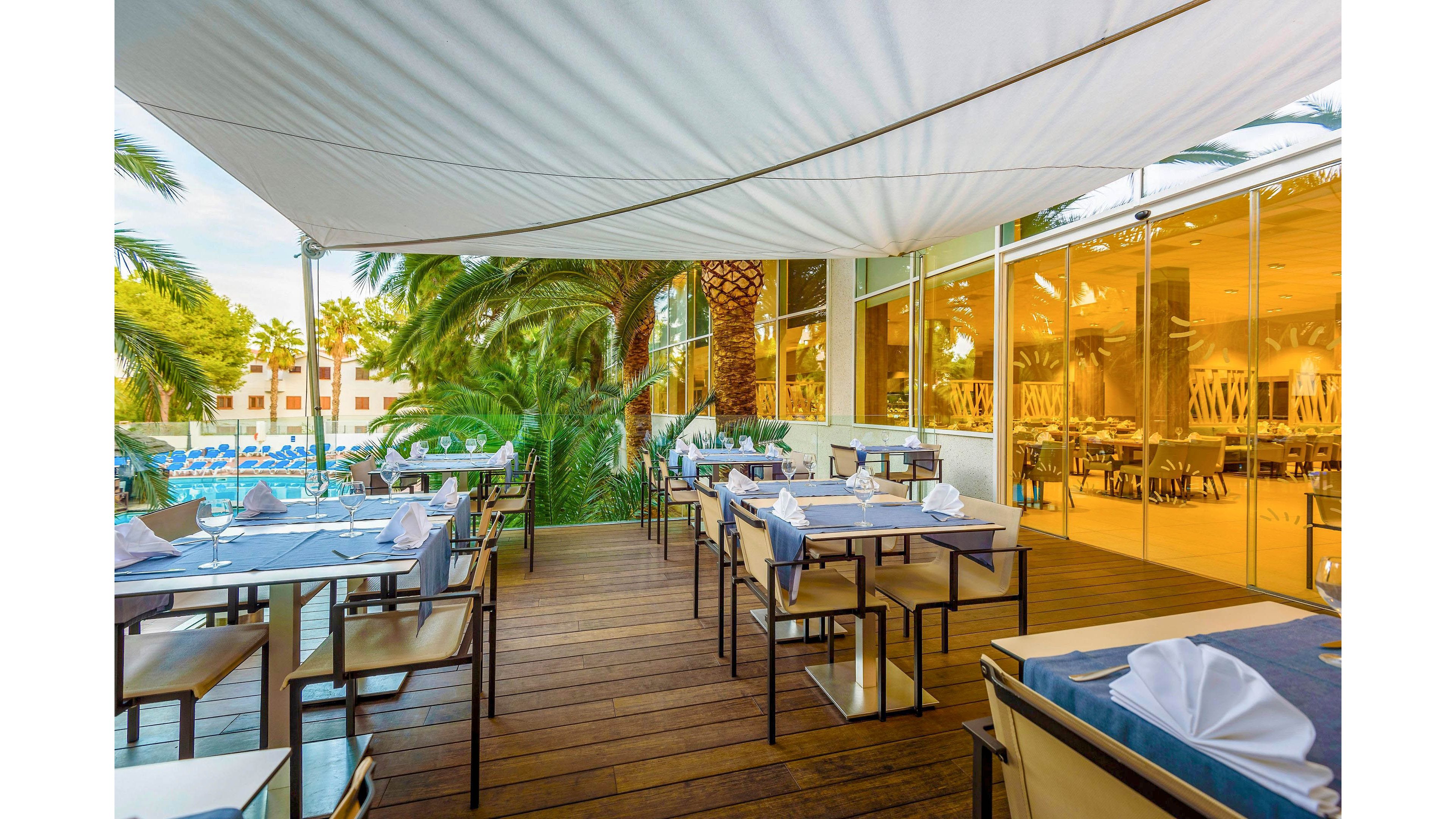 An elegant outdoor restaurant terrace is set with neatly arranged tables and chairs under a large white canopy. Each table is covered with blue runners and white napkins folded into decorative shapes, creating a refined dining atmosphere. The terrace overlooks a swimming pool surrounded by palm trees and resort buildings. Large glass windows reveal an indoor dining area, reflecting the warm ambient lighting of the restaurant.