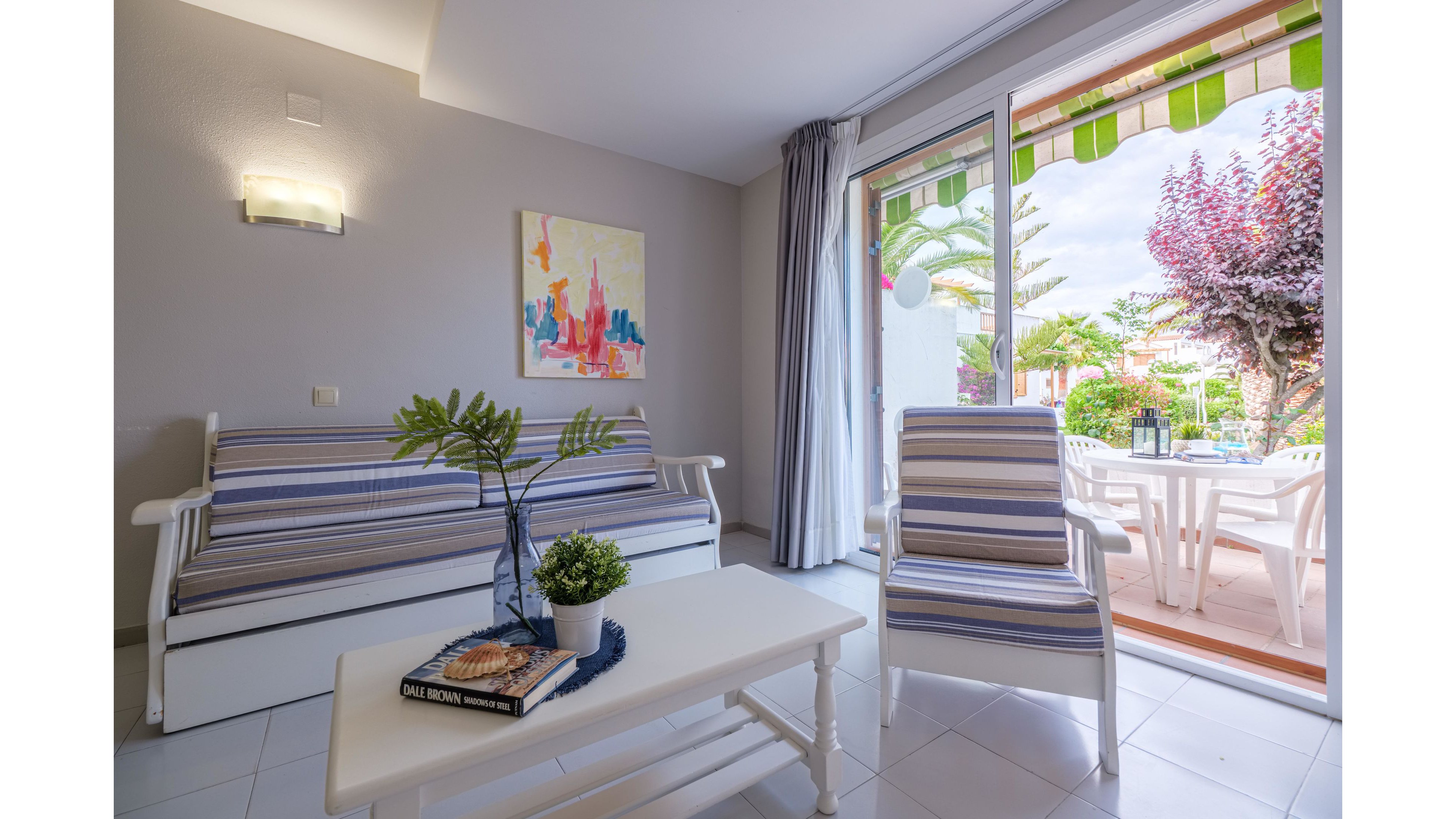 A modern living room features a white sofa and matching armchair with blue and white striped cushions. A simple white coffee table holds a decorative plant and a book, while colorful artwork hangs on the wall. Large glass doors open to a sunny terrace with white outdoor furniture and lush greenery. The space feels airy and inviting, with soft natural light and a relaxed holiday atmosphere.