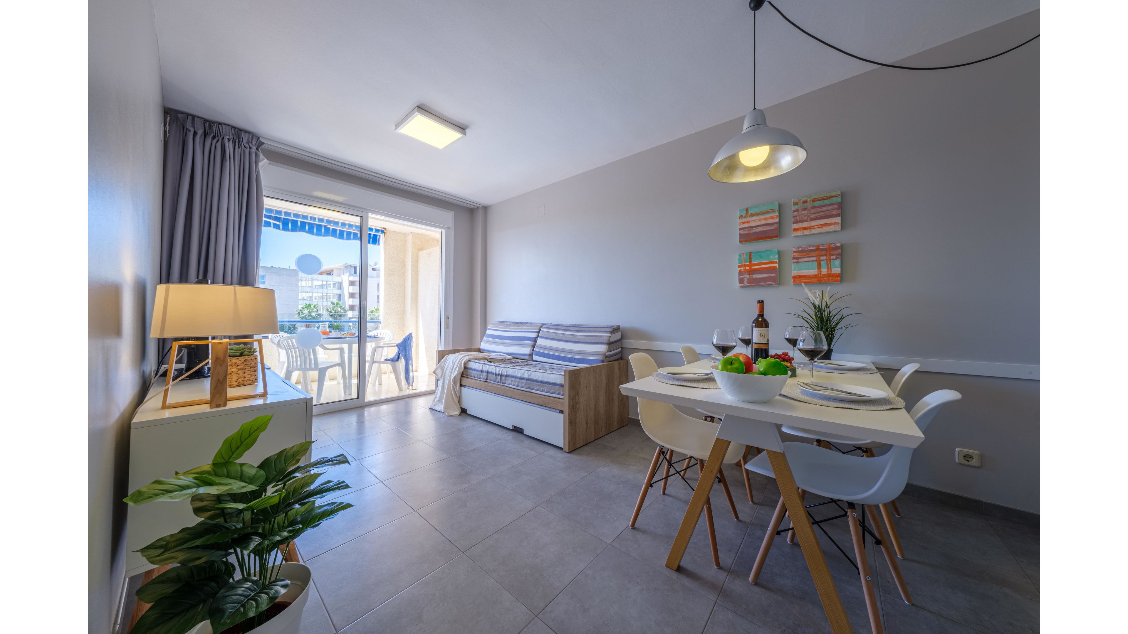 A bright modern apartment features an open-plan dining and living area with large sliding glass doors leading to a balcony. A white dining table is set with plates, glasses, and a bottle of wine, surrounded by contemporary chairs. The living space includes a compact sofa, sideboard with a lamp, indoor plants, and colorful wall art. Natural light fills the room, highlighting the clean lines and minimalist decor.