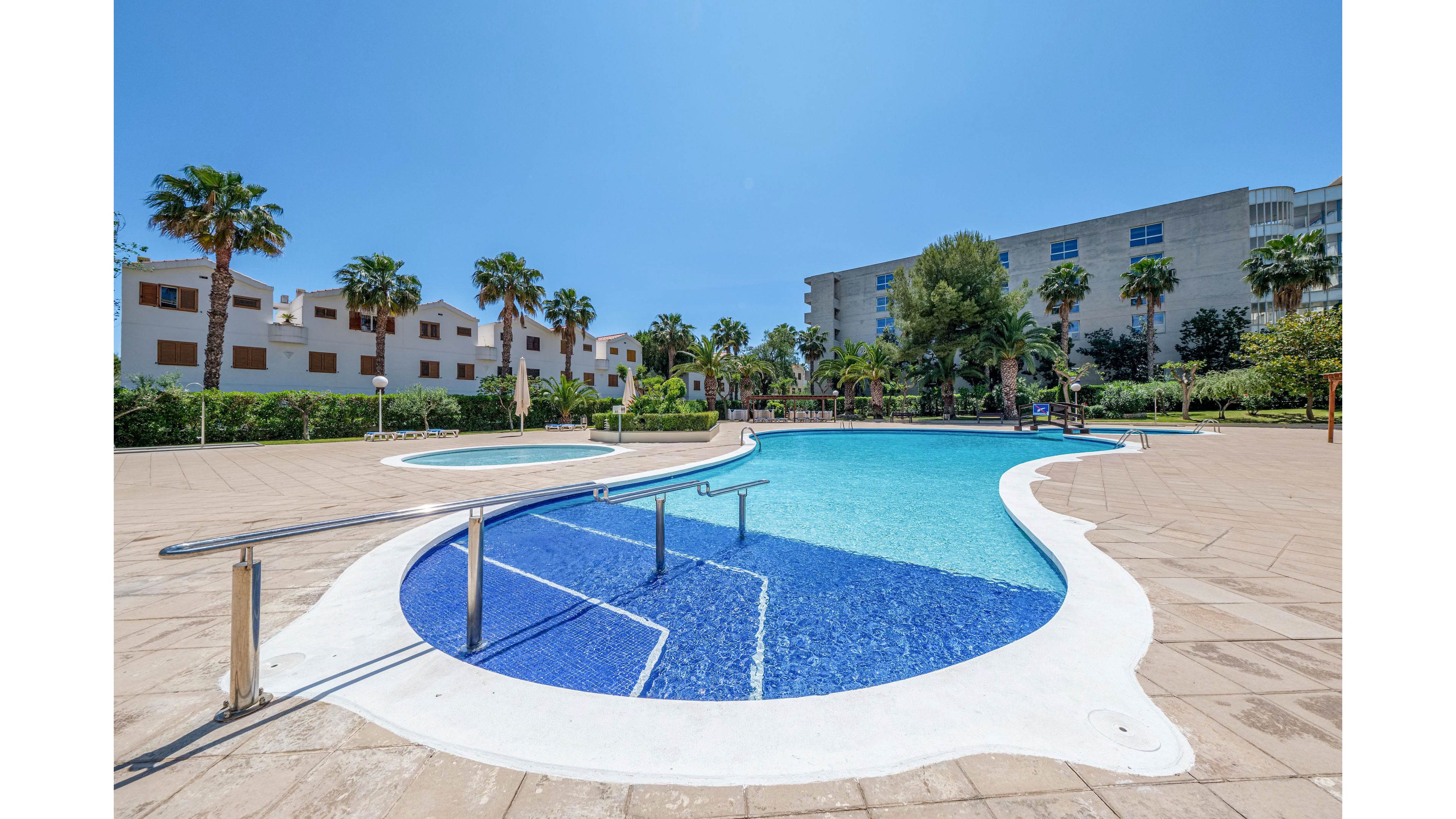 A bright outdoor swimming pool curves through a spacious resort courtyard under a clear blue sky. Palm trees and manicured greenery surround the pool, with residential-style buildings in the background. A metal handrail leads into the shallow end, and a smaller circular pool sits nearby. The scene suggests a warm, relaxing vacation atmosphere in a modern holiday complex.