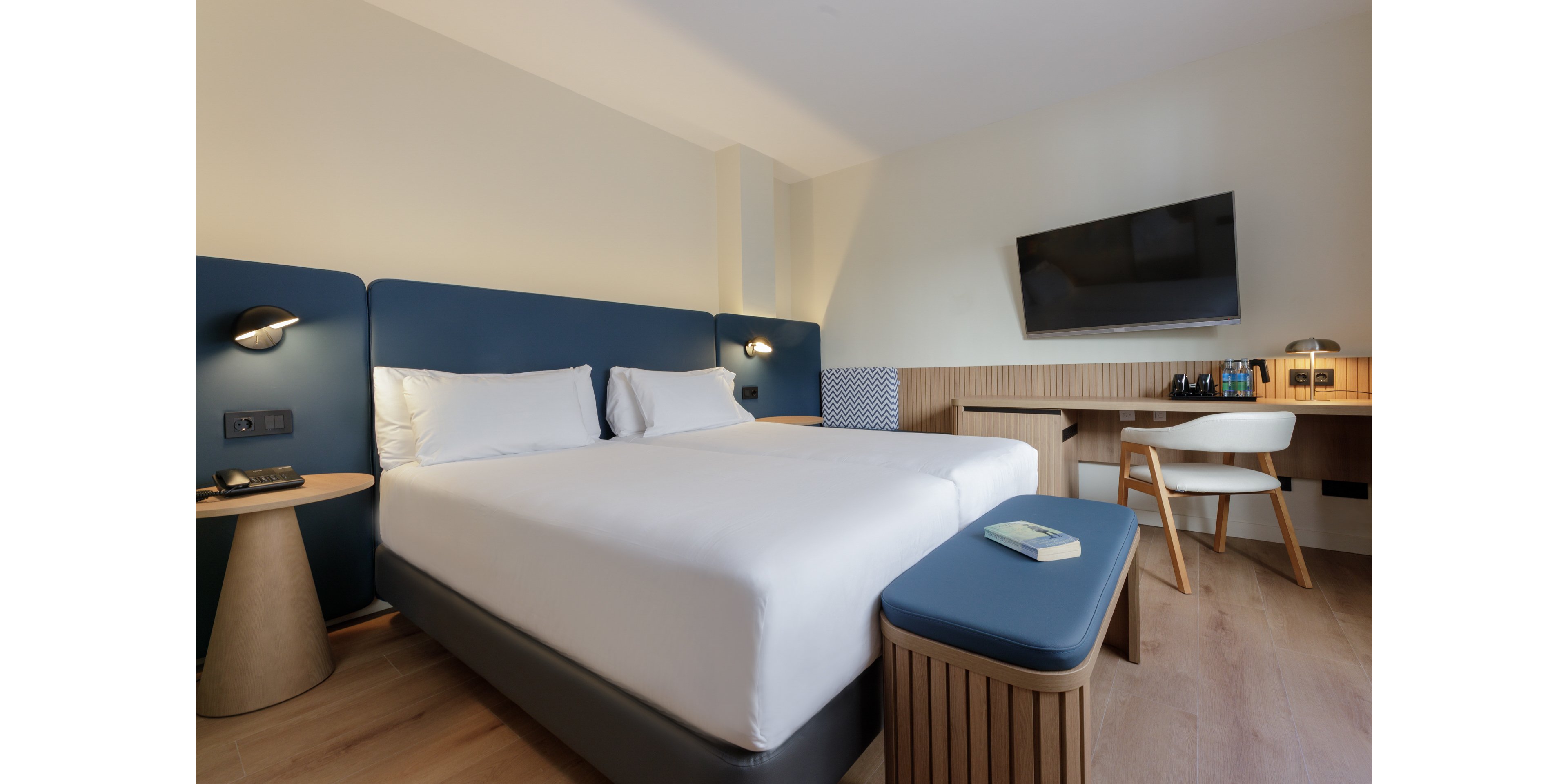 Contemporary hotel room featuring a large double bed with crisp white linens and blue upholstered headboard. A wall-mounted flat-screen TV faces the bed, and a wooden desk with a white chair sits along the side wall. A cushioned blue bench with a book on top stands at the foot of the bed, complemented by matching bedside tables and lamps. The space is bright and minimalist, with light wood flooring and neutral walls creating a calm, inviting atmosphere.