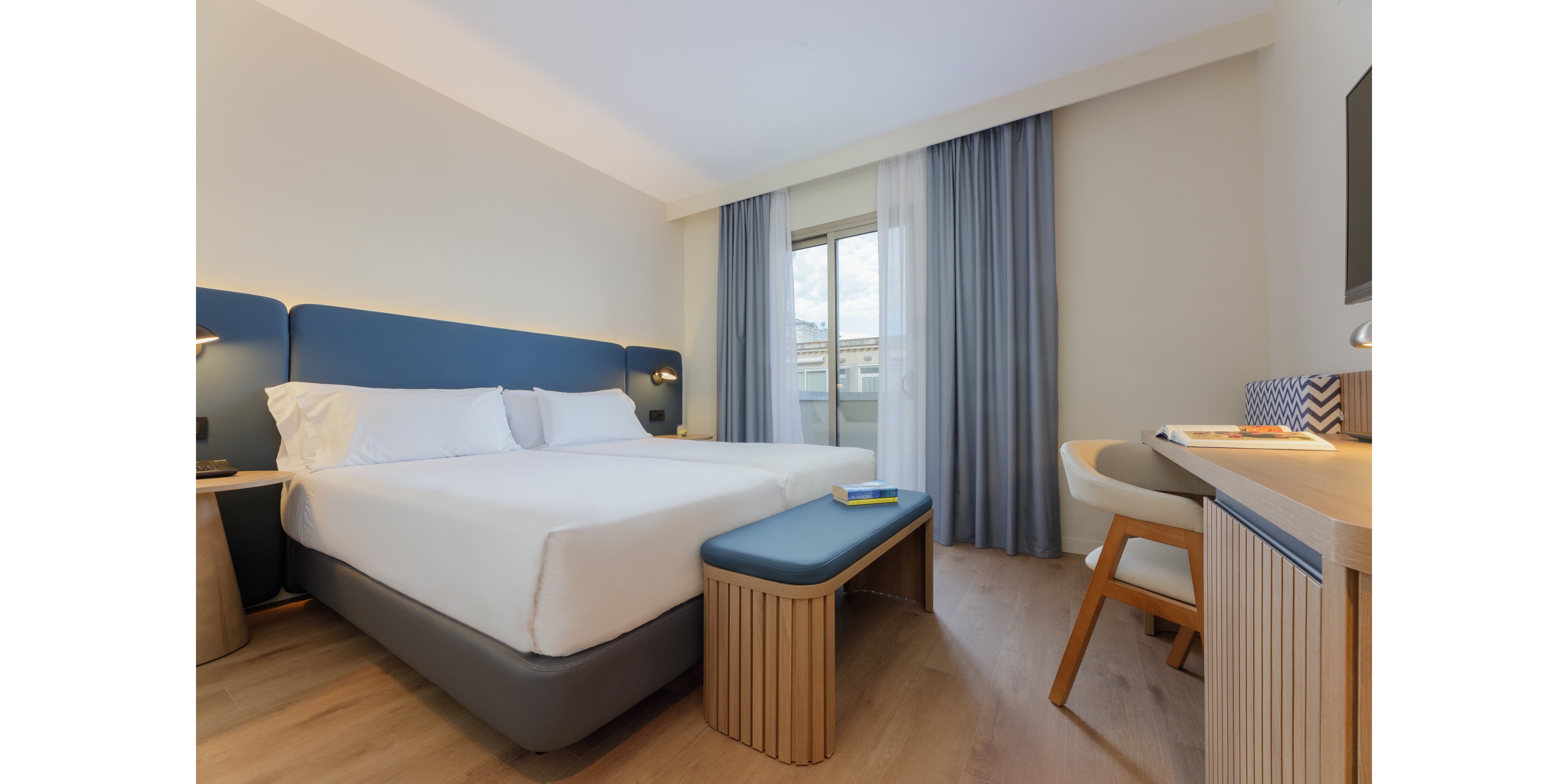 Contemporary hotel room featuring a neatly made double bed with white linens and blue upholstered headboard. The space includes a small bench at the foot of the bed and a wooden desk with a chair on the right side. Light wood flooring and soft blue and white curtains frame a window leading to a balcony with an urban view. The room is bright, minimal, and designed for comfortable short stays or business travel.