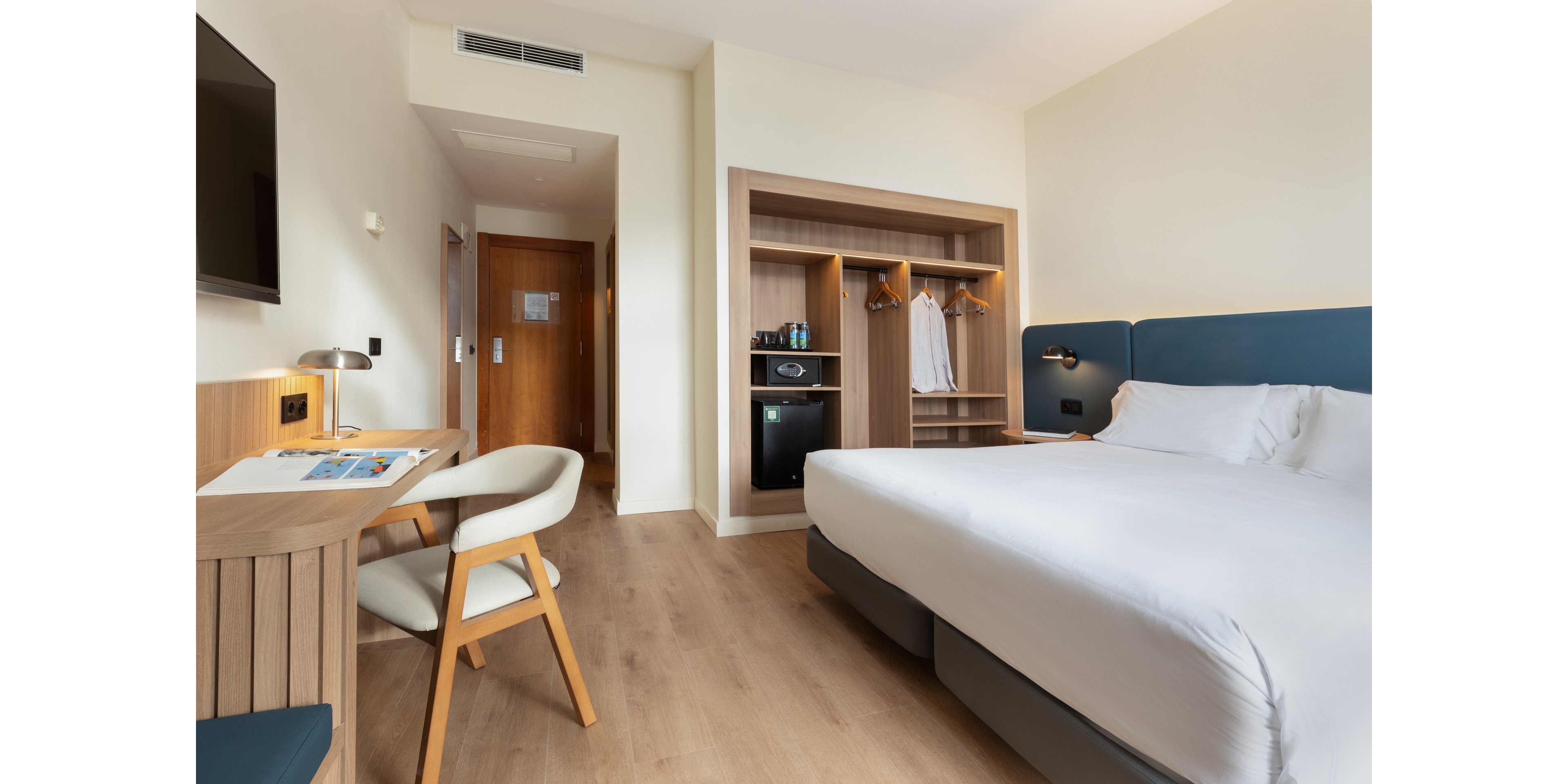 A contemporary hotel room features a neatly made double bed with white linens and a blue upholstered headboard. Opposite the bed, a wooden desk with a chair and reading lamp sits beneath a wall-mounted television. An open wardrobe unit reveals hanging clothes, shelves, and a small refrigerator, while a wooden door leads to the hallway or bathroom area. The room has light-colored walls and flooring, creating a bright, clean, and minimalist atmosphere.