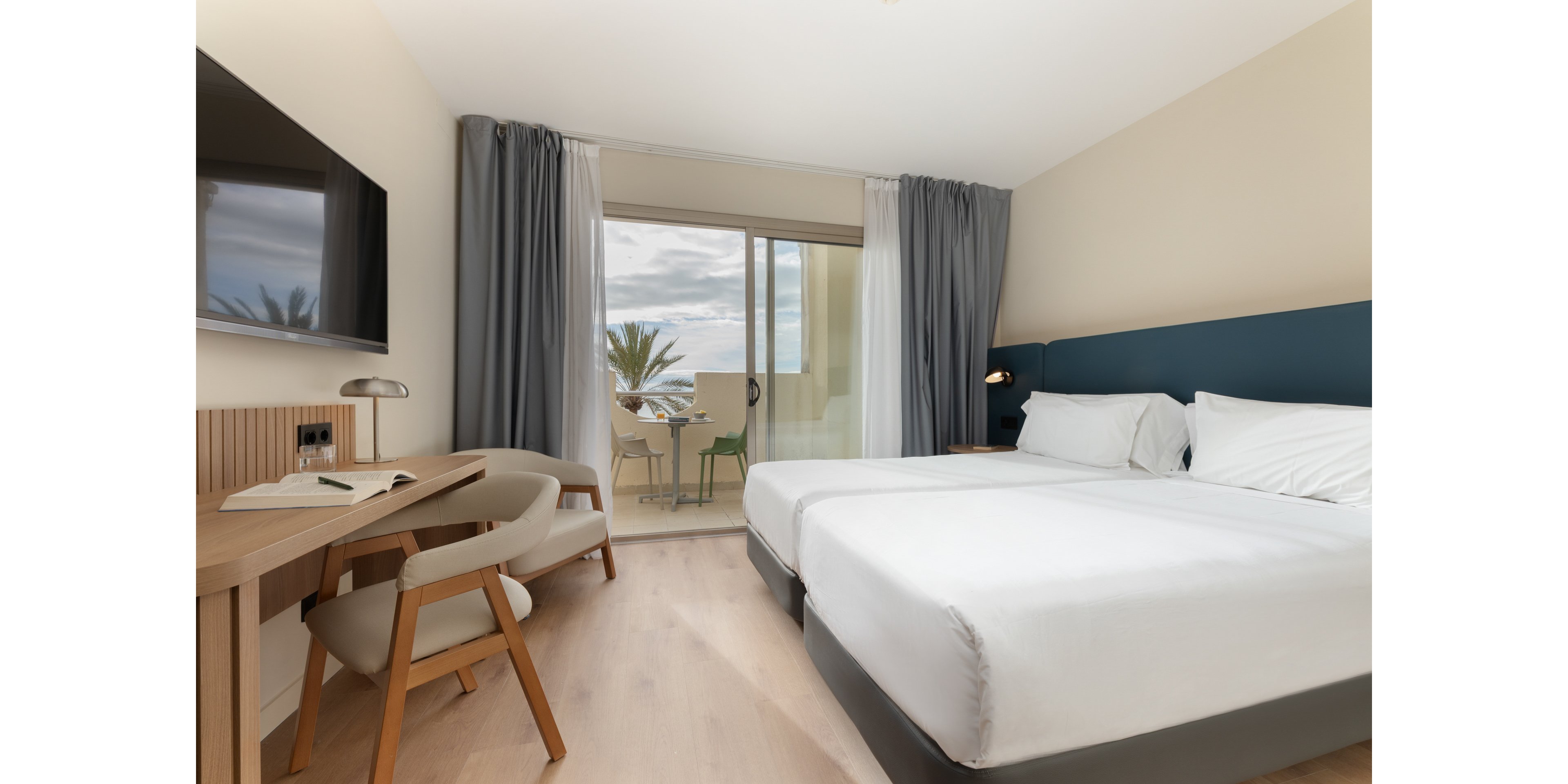 A contemporary hotel room features two neatly made single beds with white linens and a padded headboard. A wooden desk with a chair, open notebook, and lamp sits opposite a wall-mounted television. Large sliding glass doors lead to a balcony with outdoor chairs and a table, revealing palm trees and a cloudy sky. Neutral walls, light wood flooring, and soft gray and white curtains create a calm, minimalist atmosphere.