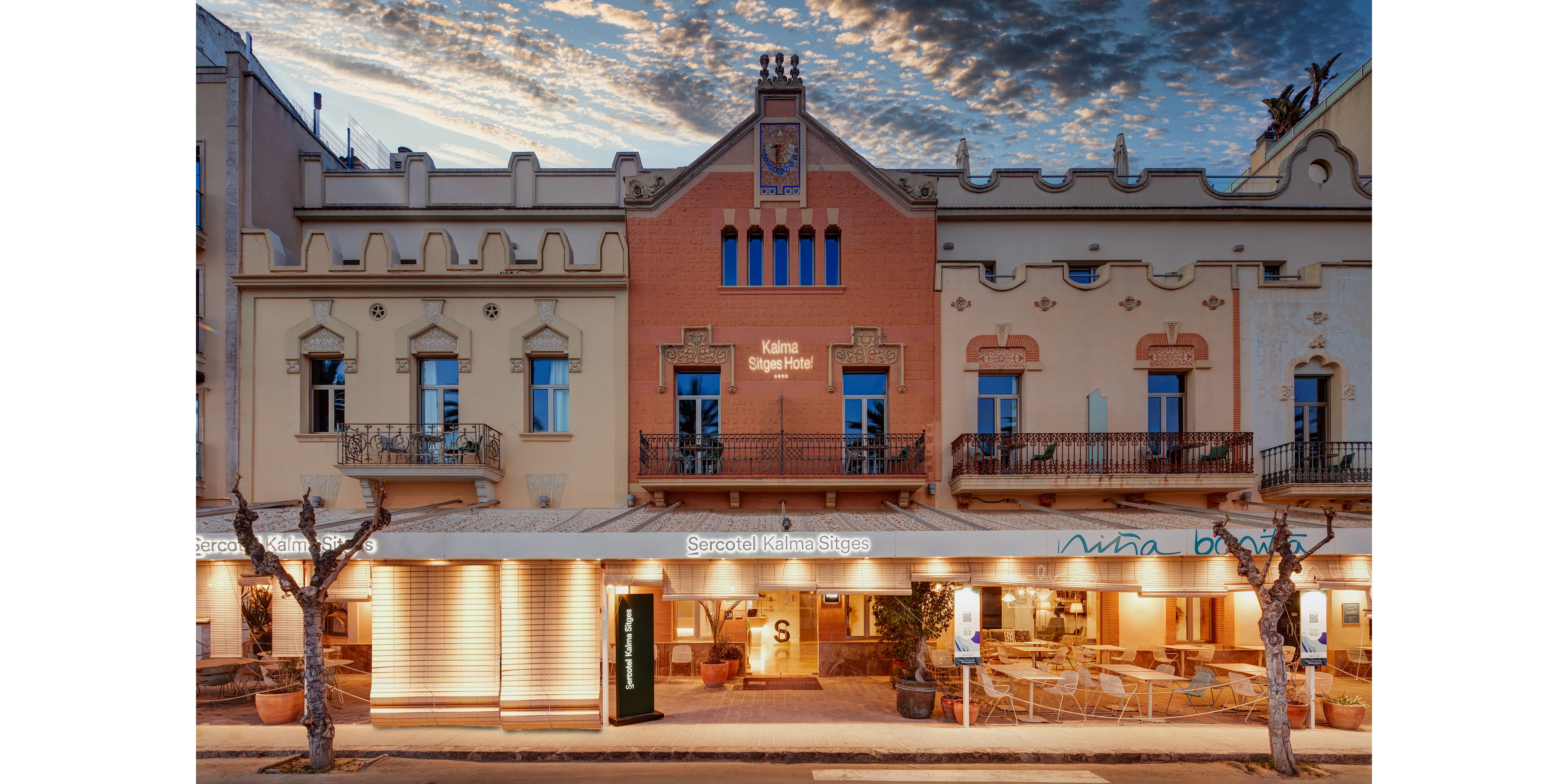 A historic hotel building with ornate architectural details is photographed from the street at dusk. Warm lights illuminate the ground-floor terrace and entrance, highlighting outdoor seating and signage. The upper floors feature balconies, arched windows, and decorative trim against a softly lit evening sky with scattered clouds. Bare trees line the sidewalk in front, framing the inviting exterior of the hotel.