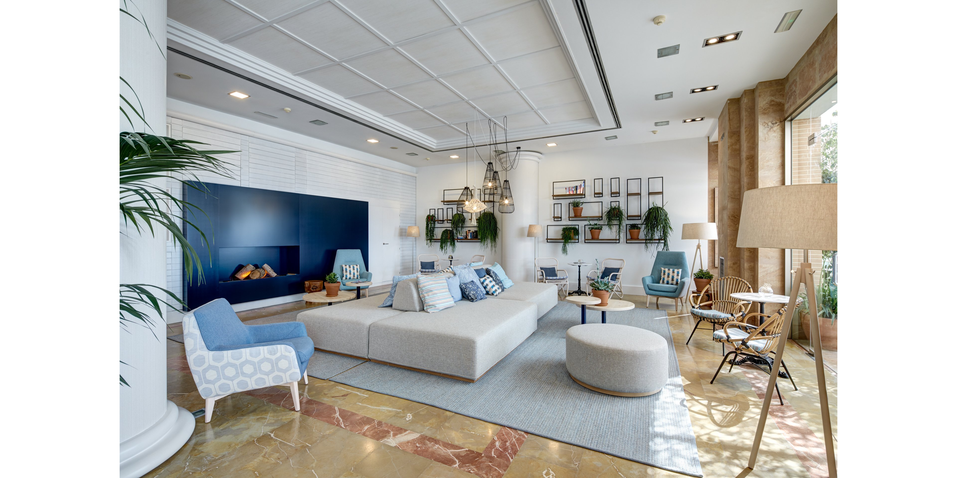 A spacious contemporary lounge features a large modular sofa with assorted cushions as the central focal point. The room includes modern armchairs, round ottomans, and small side tables arranged on a light area rug. A sleek built-in wall unit with a blue accent panel and decorative fireplace adds visual interest. Large windows bring in natural light, while hanging pendant lights, indoor plants, and open shelving create a relaxed, welcoming atmosphere.
