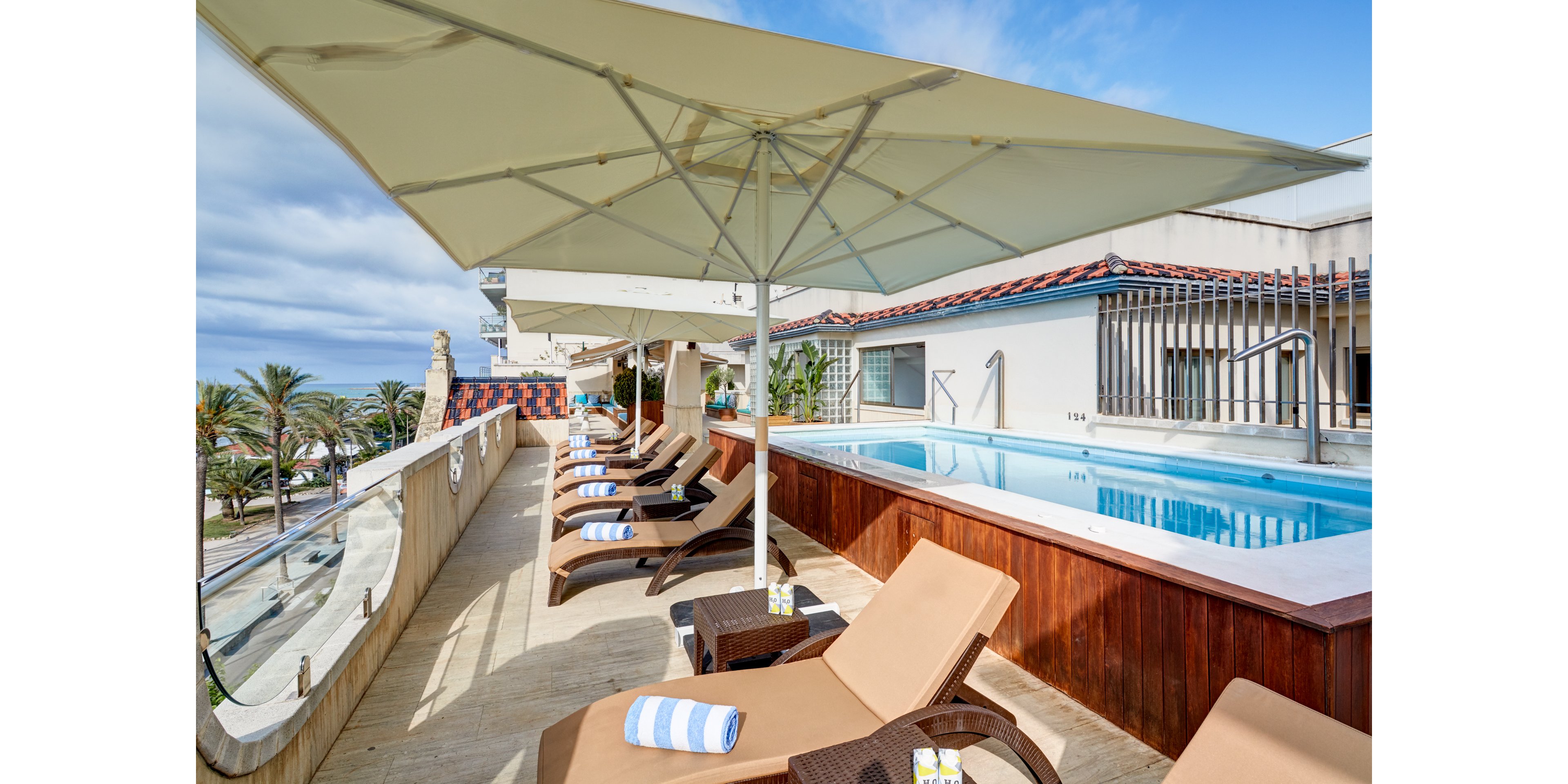 A rooftop terrace features a narrow elevated swimming pool lined with wooden paneling. Several tan sun loungers with striped towels are arranged under large white umbrellas. Small side tables hold drinks, suggesting a relaxing vacation atmosphere. In the background, palm trees, buildings, and a partly cloudy sky complete the coastal resort setting.