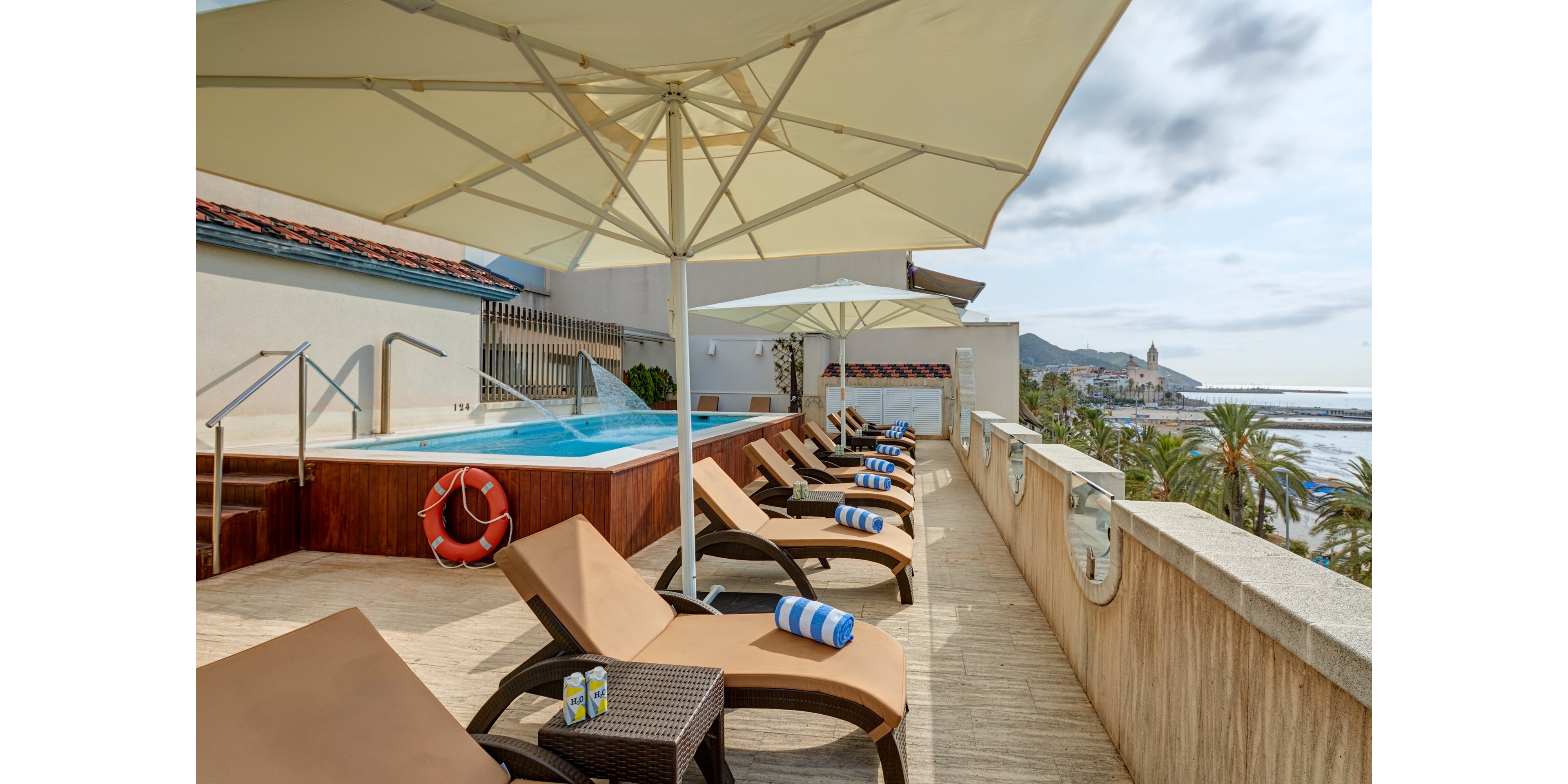 A rooftop terrace features several brown sun loungers arranged beside a small elevated pool. Large white umbrellas provide shade over the seating area, where striped blue and white towels are neatly rolled on the chairs. A bright orange lifebuoy hangs near the pool steps, and two beverage cans rest on a side table. In the background, palm trees, coastal buildings, and the sea stretch toward the horizon under a partly cloudy sky.