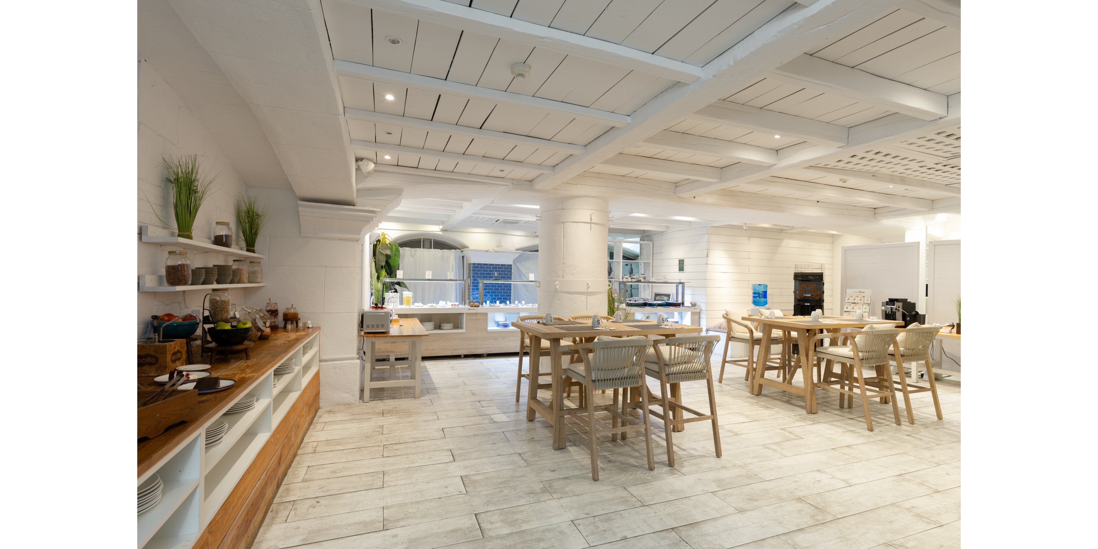 Spacious cafeteria interior featuring light wooden tables and woven chairs is shown in a bright, open-plan setting. White walls, ceiling beams, and tiled flooring create an airy, contemporary atmosphere. Open shelving with dishes and decor lines one side, while a buffet or service counter is visible in the background. Soft, even lighting and neutral tones emphasize a clean, welcoming dining environment.