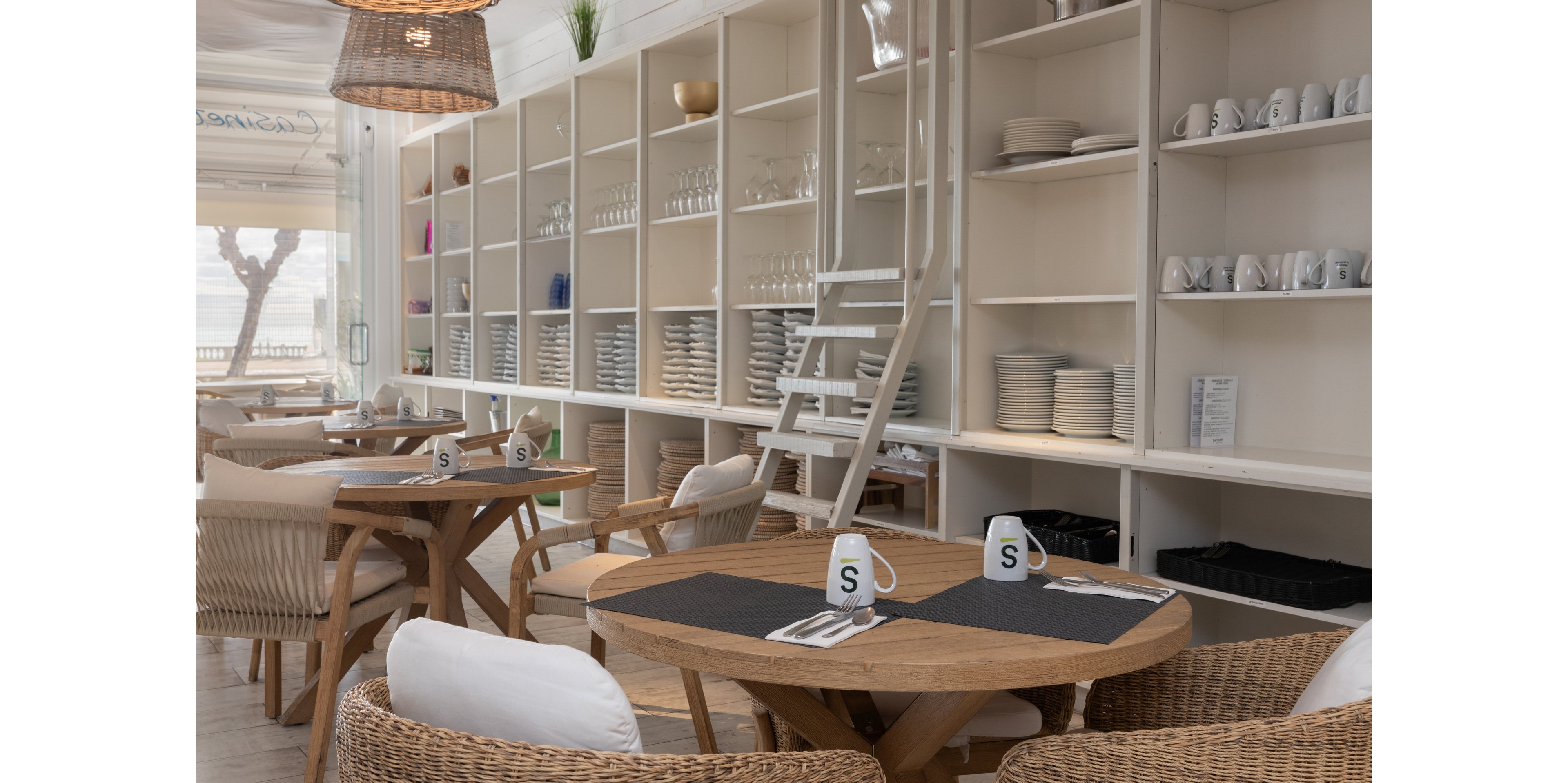 A bright, modern café interior features round wooden tables surrounded by wicker chairs with white cushions. Open shelving along the wall is neatly stacked with plates, bowls, and glassware, accessed by a white ladder. Two white mugs with a bold letter S sit on black placemats atop the tables, adding a simple graphic accent. Natural light and neutral tones create a relaxed, coastal atmosphere ideal for casual dining or coffee breaks.