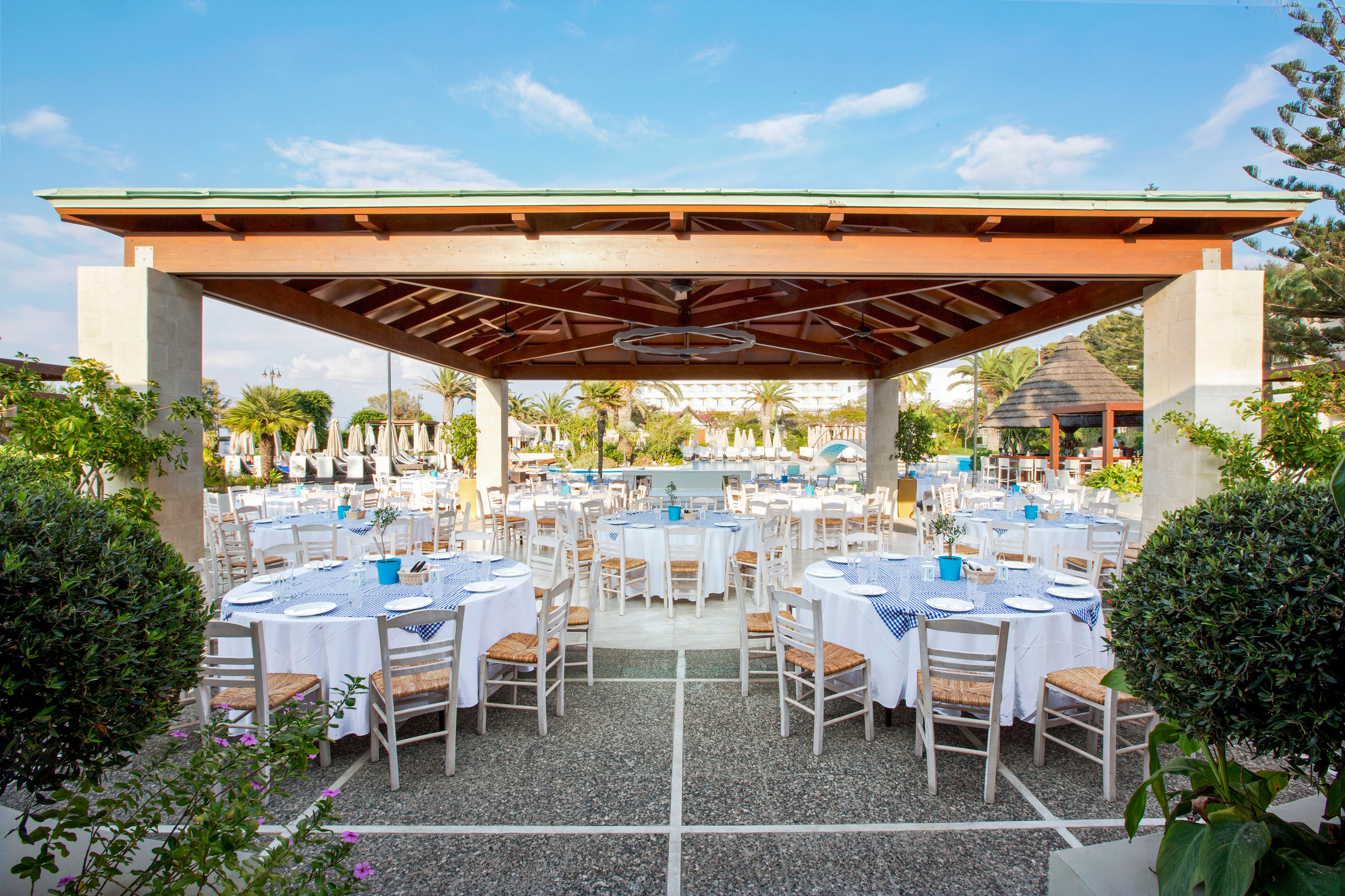 An open-air restaurant is arranged with round tables covered in white tablecloths and simple wooden chairs. The setting overlooks a swimming pool surrounded by sun loungers and umbrellas, suggesting a resort environment. Blue accents on the tables complement the clear sky and bright daylight. The scene appears ready for a formal meal or celebration in a relaxed, coastal atmosphere.