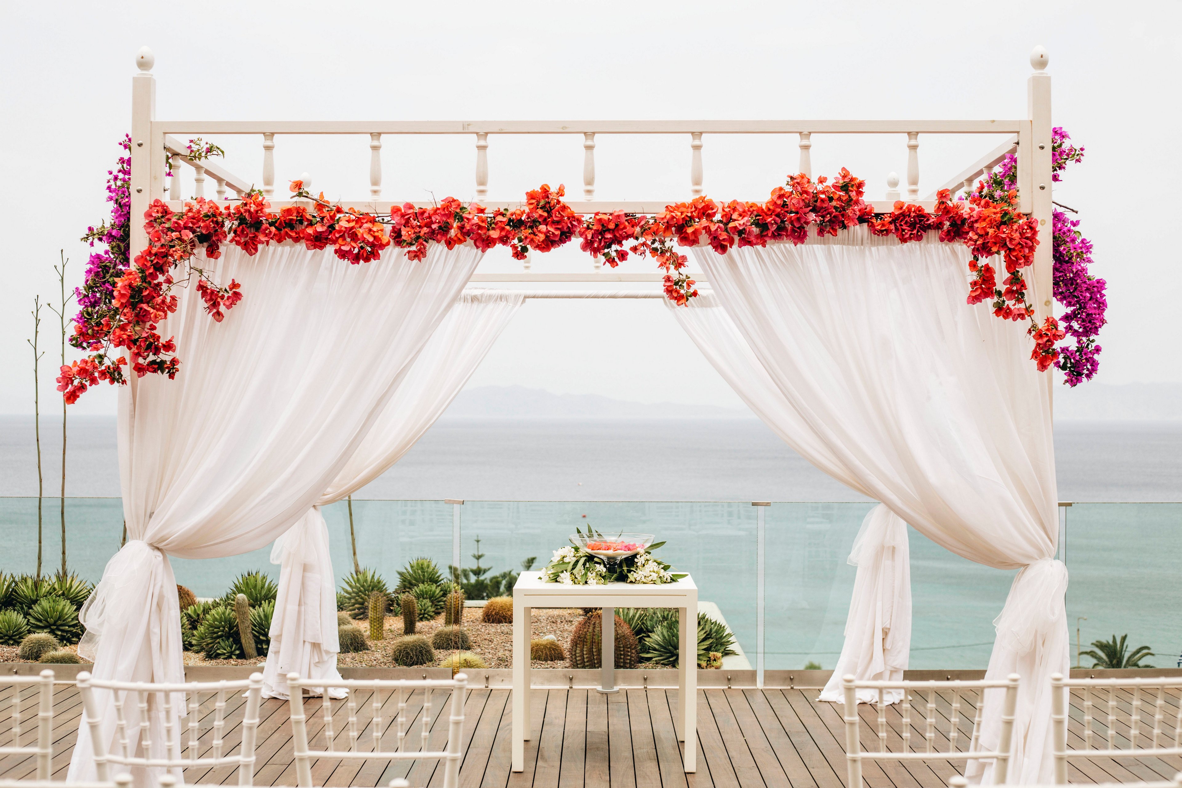 A white wedding arch decorated with flowing sheer curtains stands on a wooden deck overlooking the sea. Bright pink and red flowers cascade across the top of the structure, adding vibrant color to the soft neutral setting. White chairs are arranged in rows facing a small table with a floral arrangement beneath the arch. Succulents and coastal plants line the edge of the deck, with calm ocean water and a hazy horizon in the background.