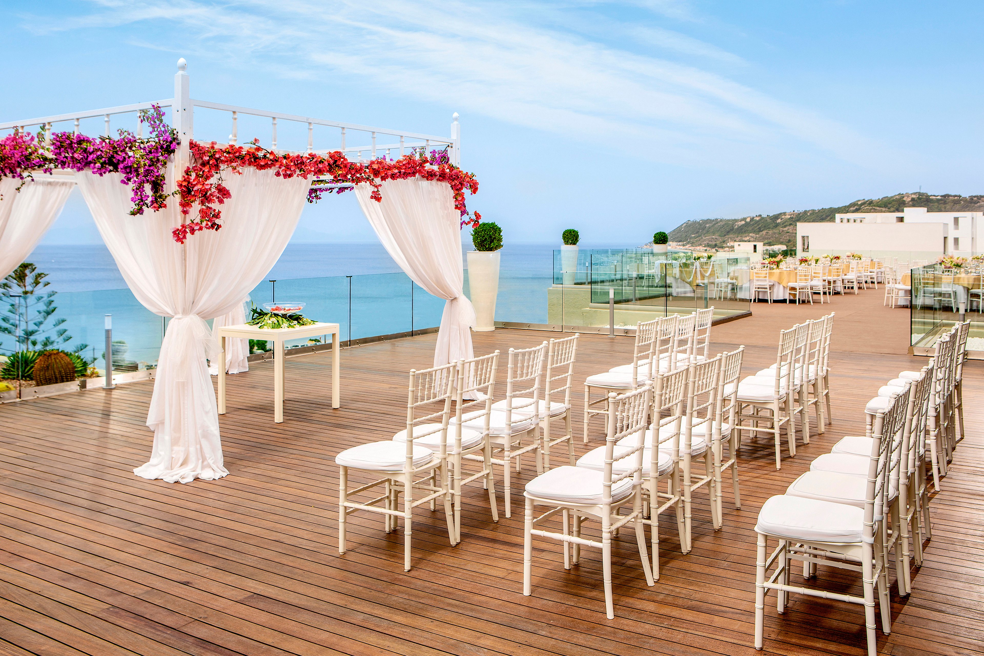 An elegant wedding ceremony setup is arranged on a wooden rooftop terrace overlooking the ocean. White chairs are lined in neat rows facing a draped canopy adorned with colorful flowers. Glass railings frame the deck, revealing a coastal landscape and blue sky in the background. Additional tables and seating are visible further along the terrace, suggesting a reception area.