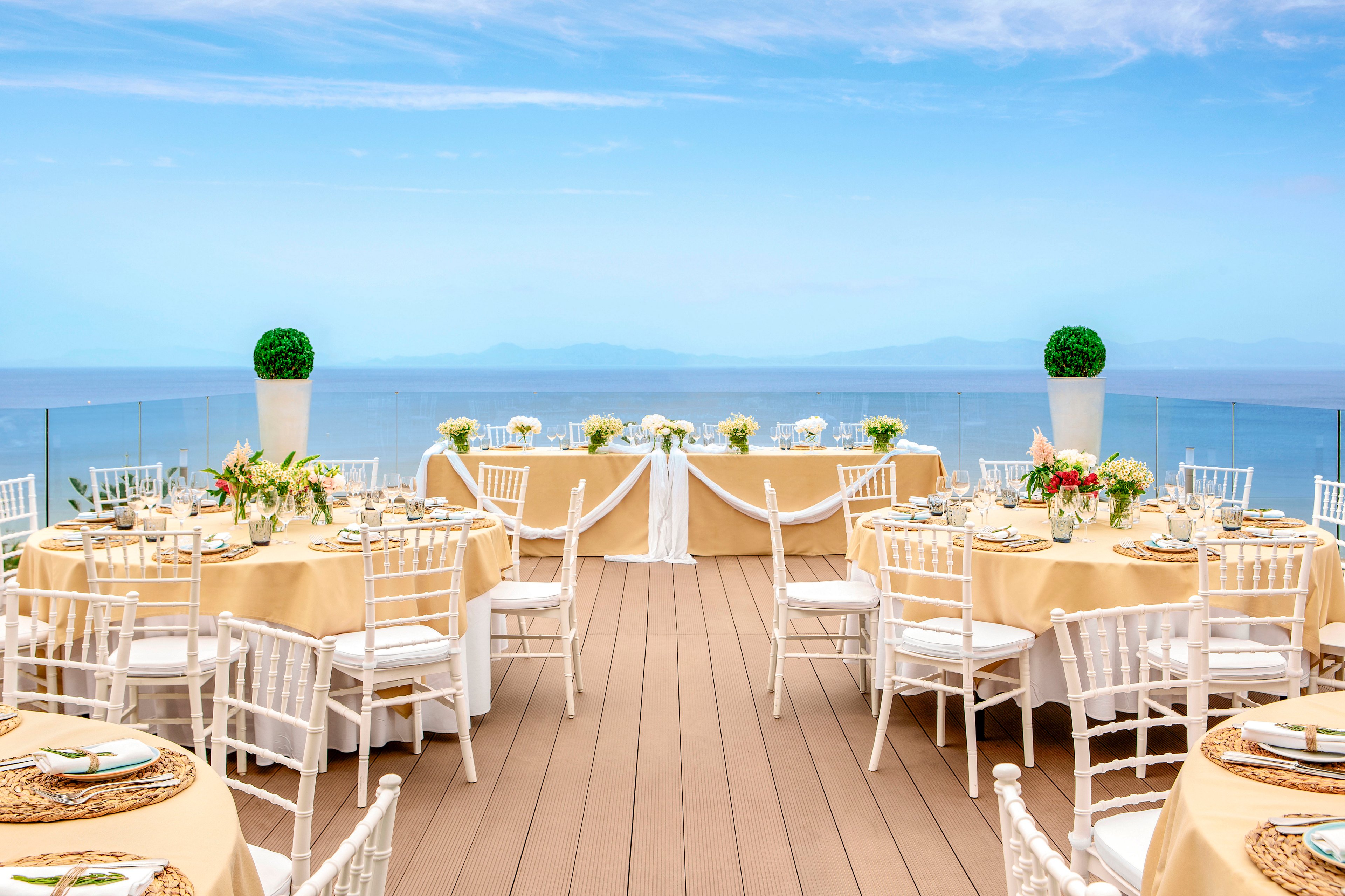 Round tables with cream-colored linens and white chairs are arranged on an open-air terrace overlooking a calm ocean. Floral centerpieces and neatly set place settings suggest a formal event, likely a wedding reception or special celebration. The clear glass railing and expansive blue sky emphasize the coastal location. Two tall white planters with manicured greenery frame the head table, adding a touch of symmetry and sophistication.