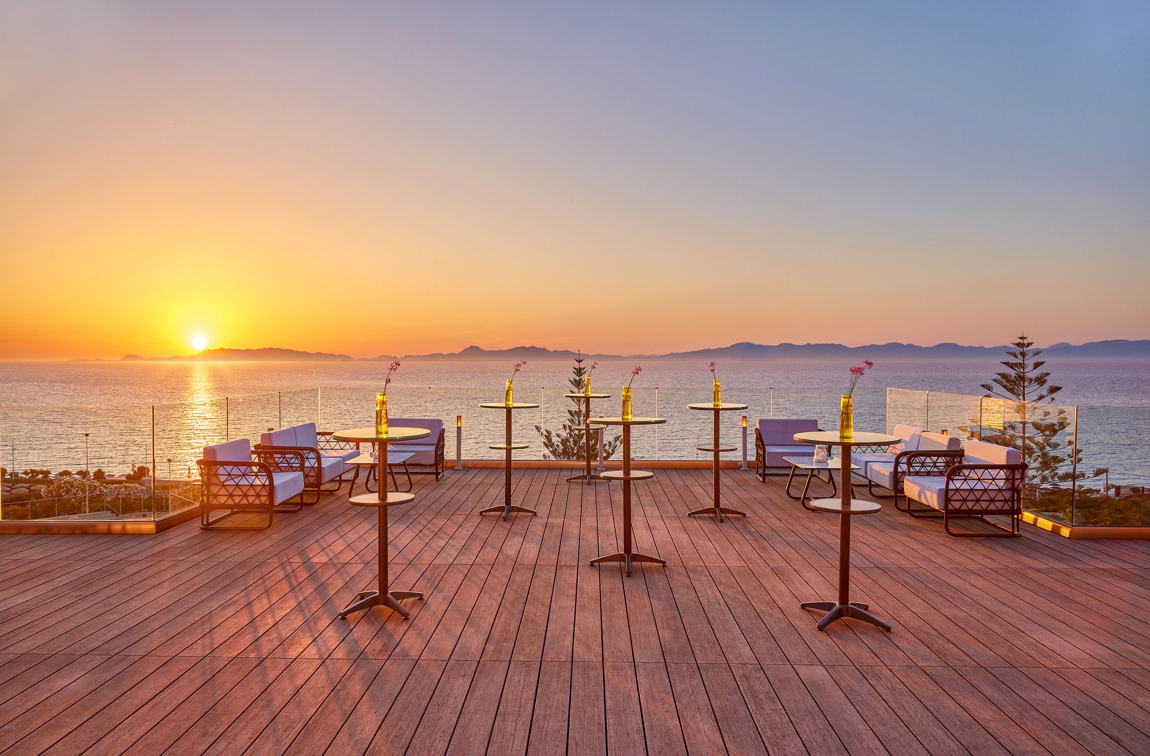 An elevated wooden terrace overlooks a calm ocean at sunset, with the sun low on the horizon casting warm golden light. Several tall cocktail tables with small vases and stools are arranged neatly across the deck, suggesting a relaxed outdoor event or lounge setting. Glass railings frame the view of the sea and distant mountains, enhancing the open, airy atmosphere. The scene feels tranquil and inviting, with soft pastel colors in the sky and reflections on the water.
