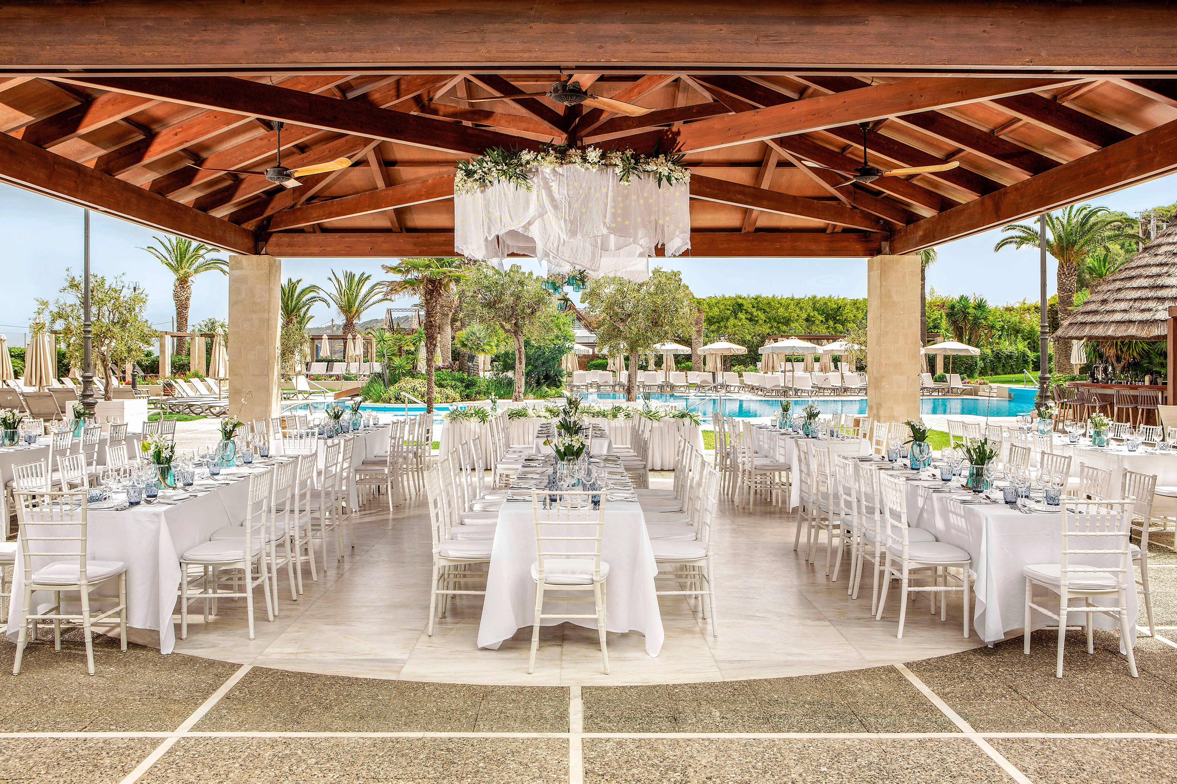 An elegant outdoor reception is set under a wooden-roof pavilion beside a large swimming pool. Long white-clothed tables with white chiavari chairs are arranged in rows, decorated with floral centerpieces and glassware. The open-air venue features stone columns, ceiling fans, and a hanging floral chandelier. In the background, palm trees, sun loungers, and resort buildings create a tropical, resort-style atmosphere.