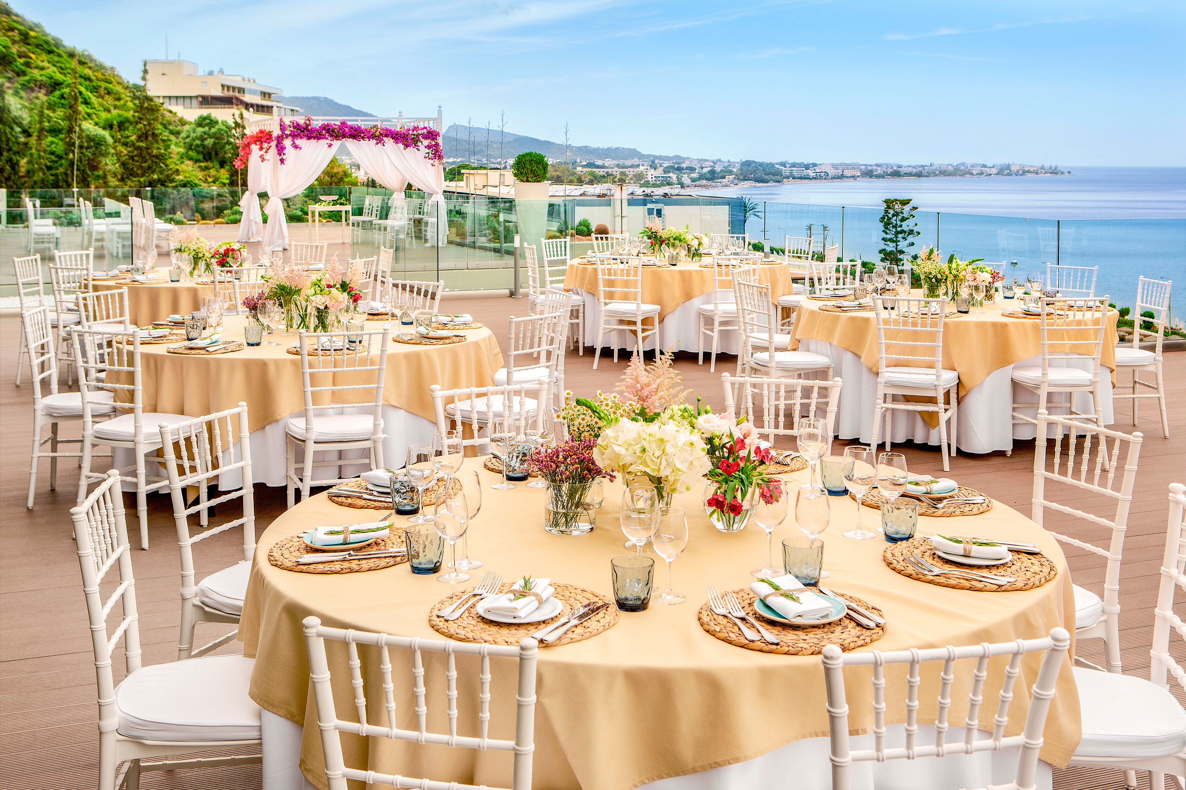 An elegant outdoor wedding reception is set on a terrace overlooking the sea. Round tables with beige tablecloths and white chiavari chairs are arranged neatly across the venue. Each table is decorated with floral centerpieces, glassware, and woven placemats. In the background, a floral arch frames the scenic coastal view under a clear blue sky.