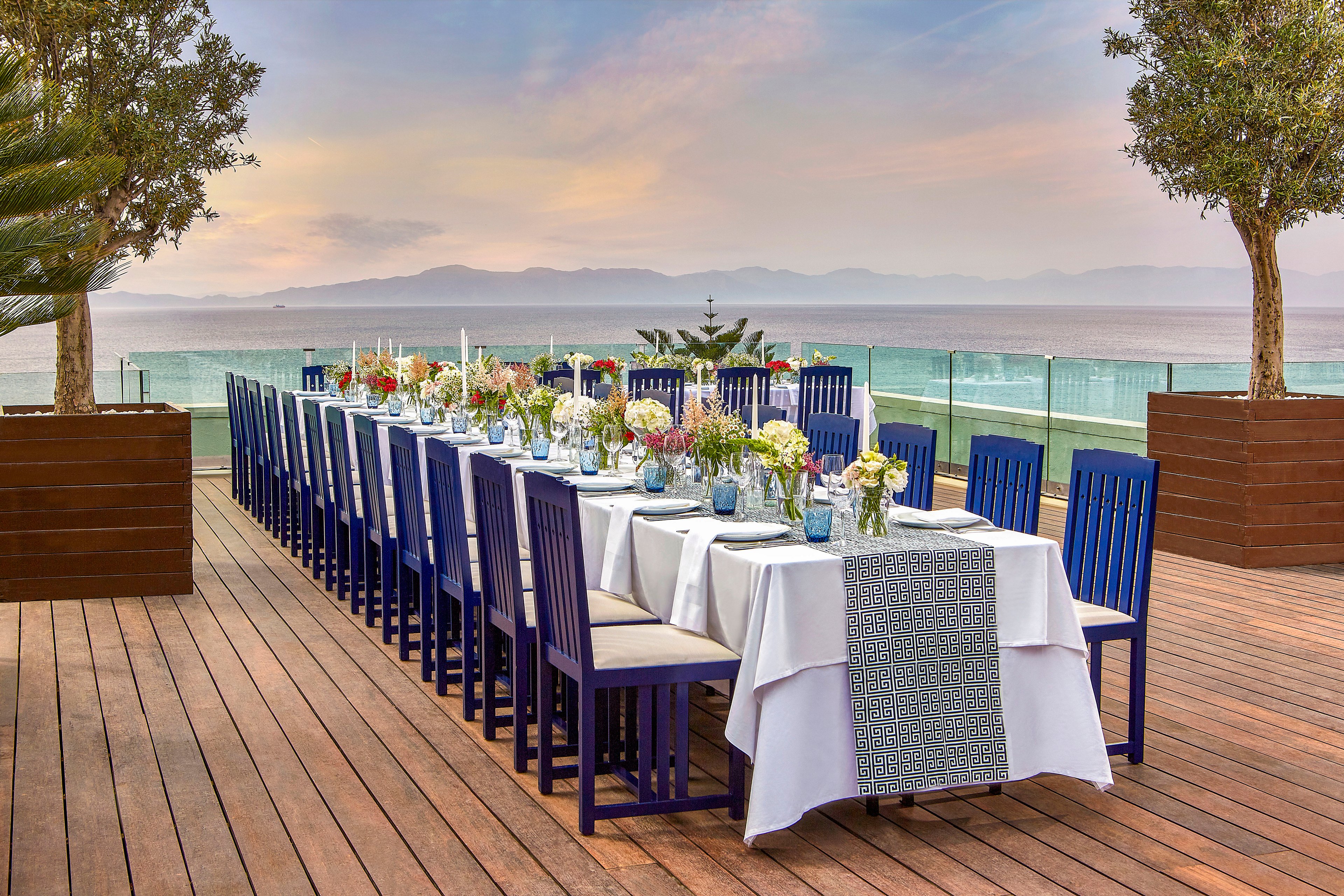 A long, elegantly set dining table is arranged on a wooden deck overlooking the ocean. Blue chairs line both sides of the table, which is covered with white linens and a patterned table runner. Glass vases filled with colorful flowers and candles decorate the tabletop, creating a festive yet refined atmosphere. The scene is framed by potted trees and a glass railing, with calm water and distant mountains under a pastel evening sky.