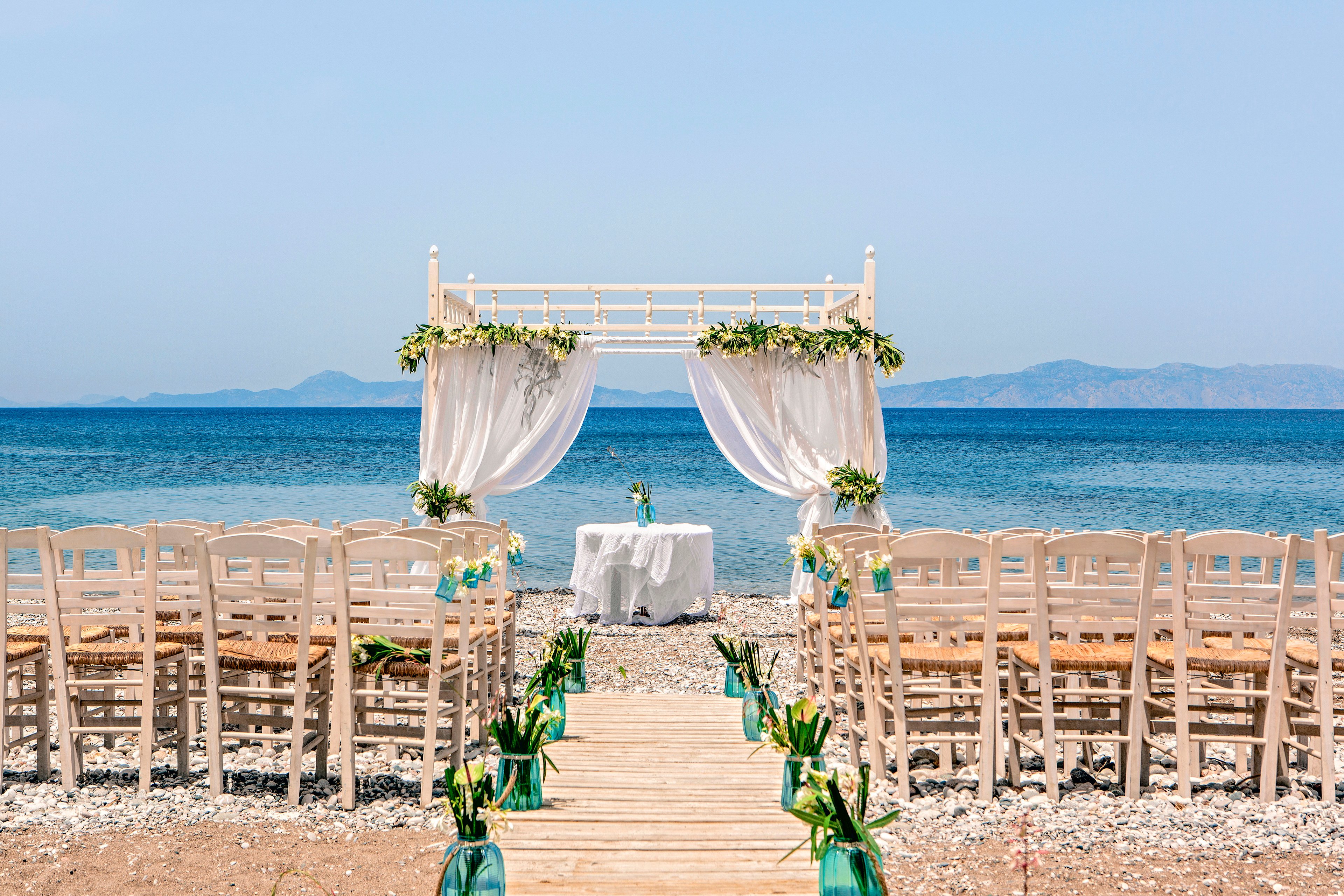 An elegant wedding ceremony setup is arranged on a pebble beach facing a calm blue sea. A wooden aisle leads to a white arch draped with sheer curtains and decorated with green foliage. Rows of white chairs line both sides of the aisle, each adorned with small floral arrangements. Glass vases with greenery flank the walkway, adding a fresh touch to the coastal scene under a clear sky.