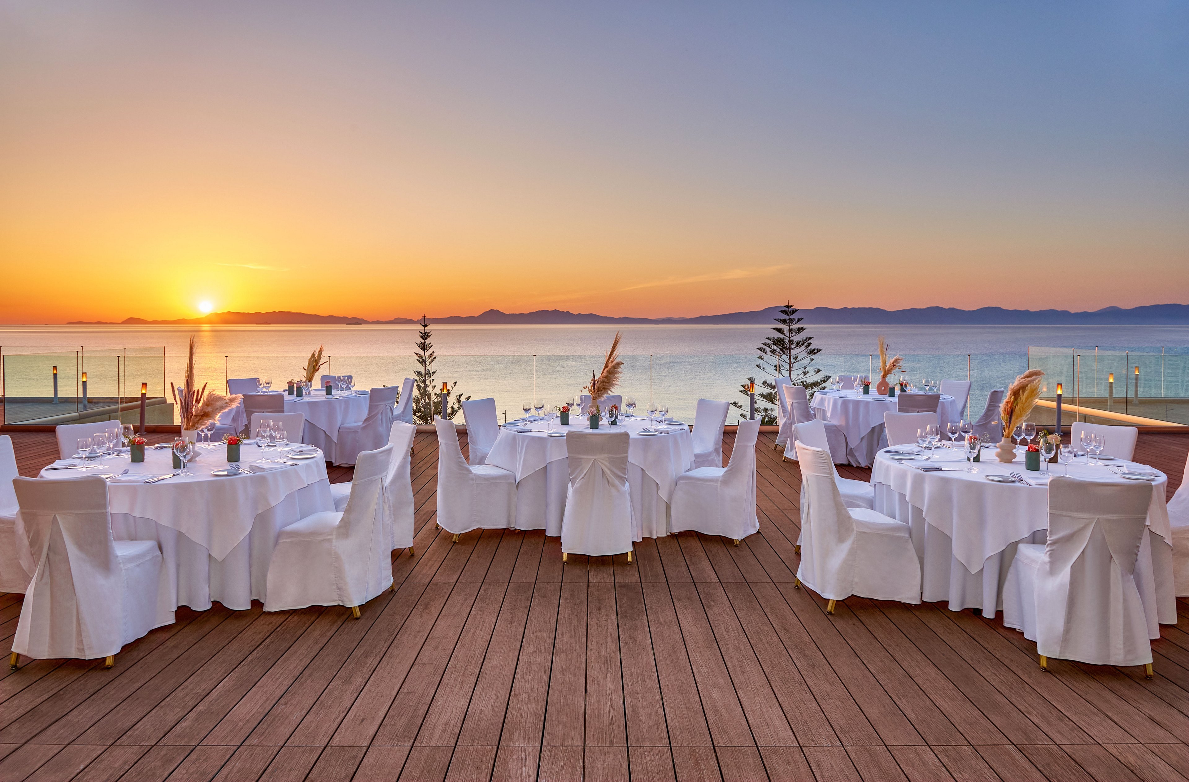 Round tables covered in white linens are arranged on a wooden terrace overlooking the sea at sunset. Each table is set with glassware, plates, and simple floral centerpieces, suggesting a formal dinner or wedding reception. The calm water and distant mountains create a serene backdrop behind the glass railing. Warm golden light from the setting sun enhances the romantic and tranquil atmosphere of the outdoor venue.