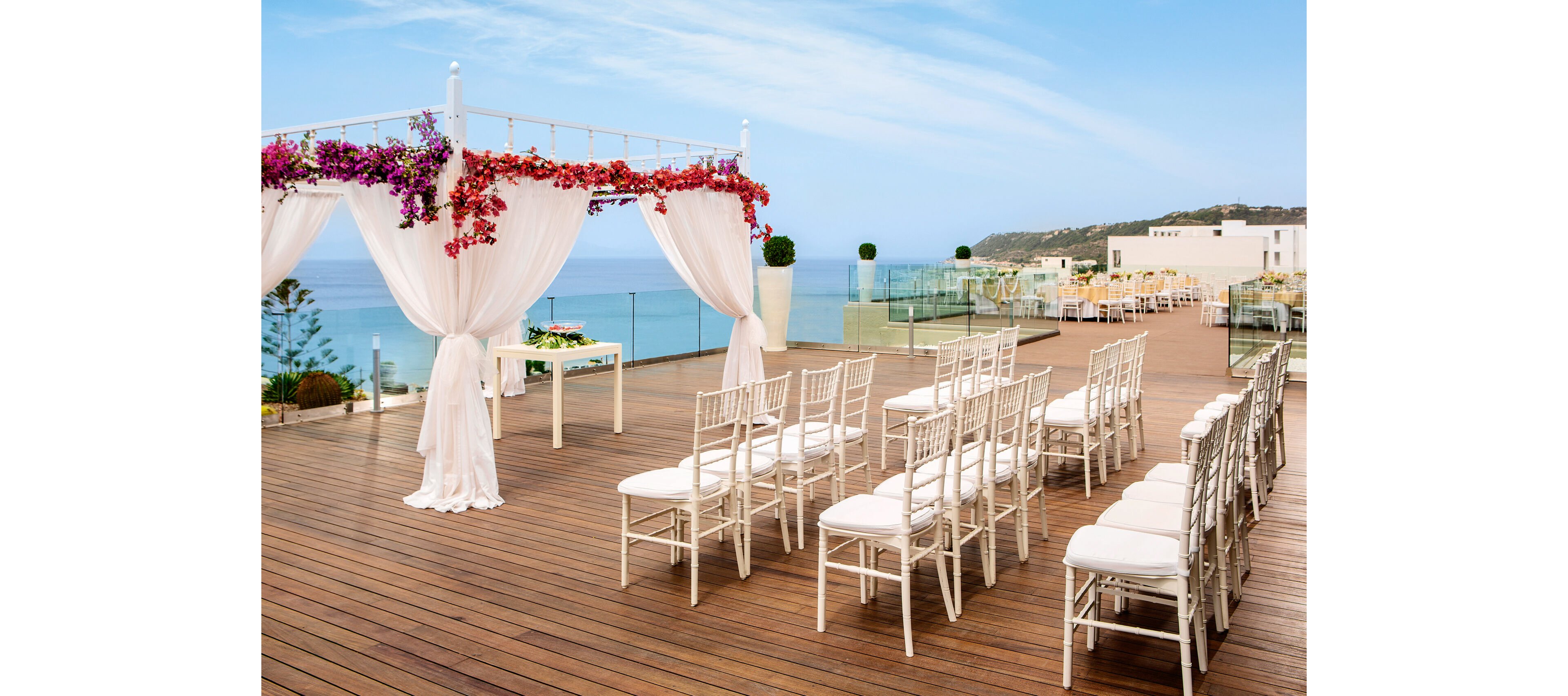 An elegant wedding ceremony setup is arranged on a wooden terrace overlooking the ocean. White chairs are lined in neat rows facing a draped canopy adorned with vibrant flowers. Glass railings frame the deck, revealing coastal cliffs and modern buildings in the background. The scene is bright and airy, suggesting a stylish outdoor event in a serene seaside location.