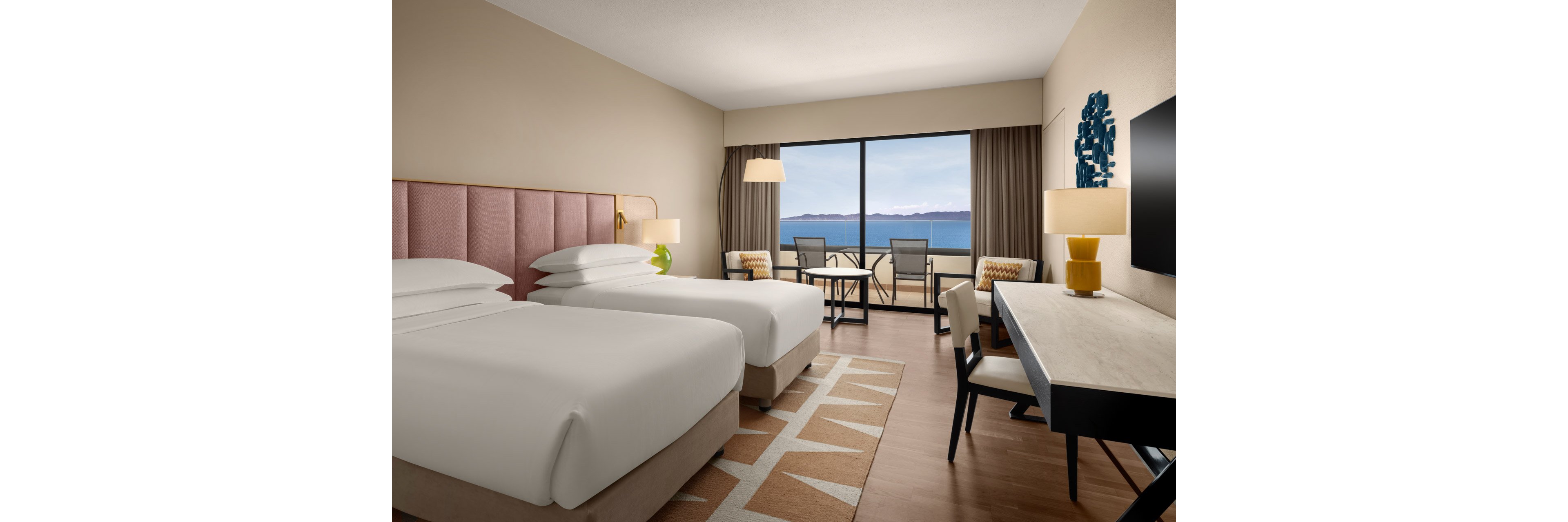 A contemporary hotel room features two neatly made twin beds with upholstered headboards. The space includes a desk with chair, a floor lamp, and patterned accent chairs around a small table. Large windows open to a balcony with seating, overlooking a calm body of water and distant mountains. Neutral tones with soft pink and beige accents create a clean, relaxing atmosphere suitable for travelers.
