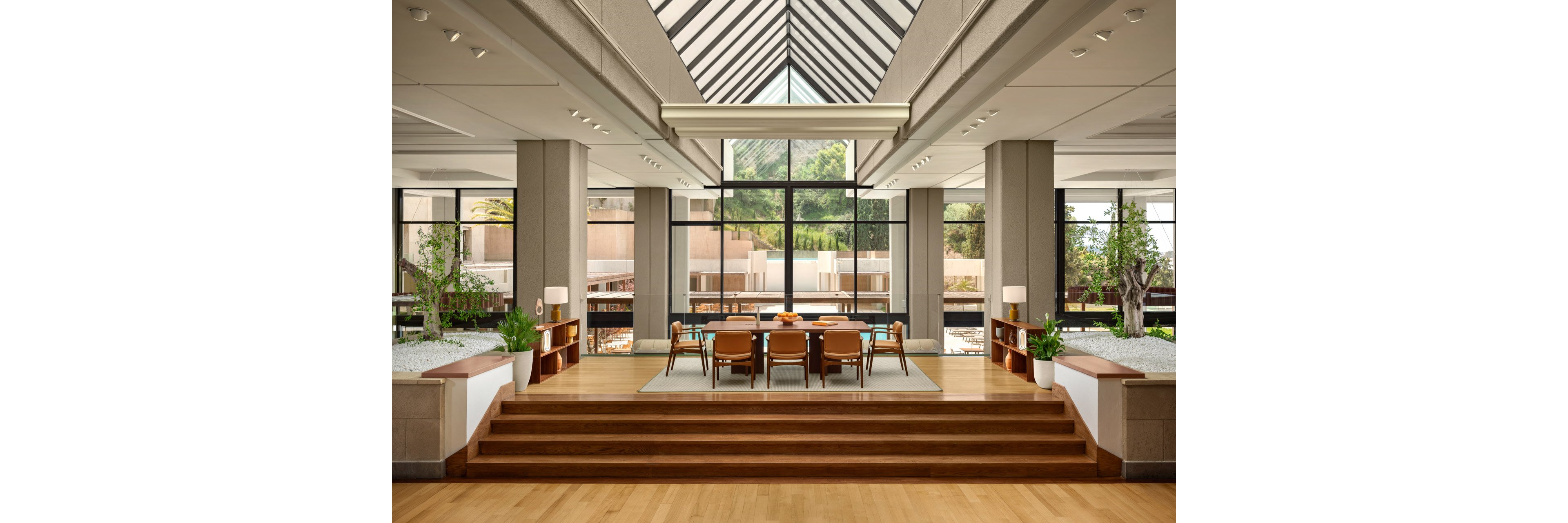 Spacious contemporary interior features a central seating area with wooden chairs around a table. The room is framed by tall columns and a dramatic glass skylight above, leading the eye toward large floor-to-ceiling windows. Natural light floods the space, highlighting warm wood flooring, neutral walls, and green plants on both sides. Outside, terraces and greenery are visible, enhancing the open, airy atmosphere.