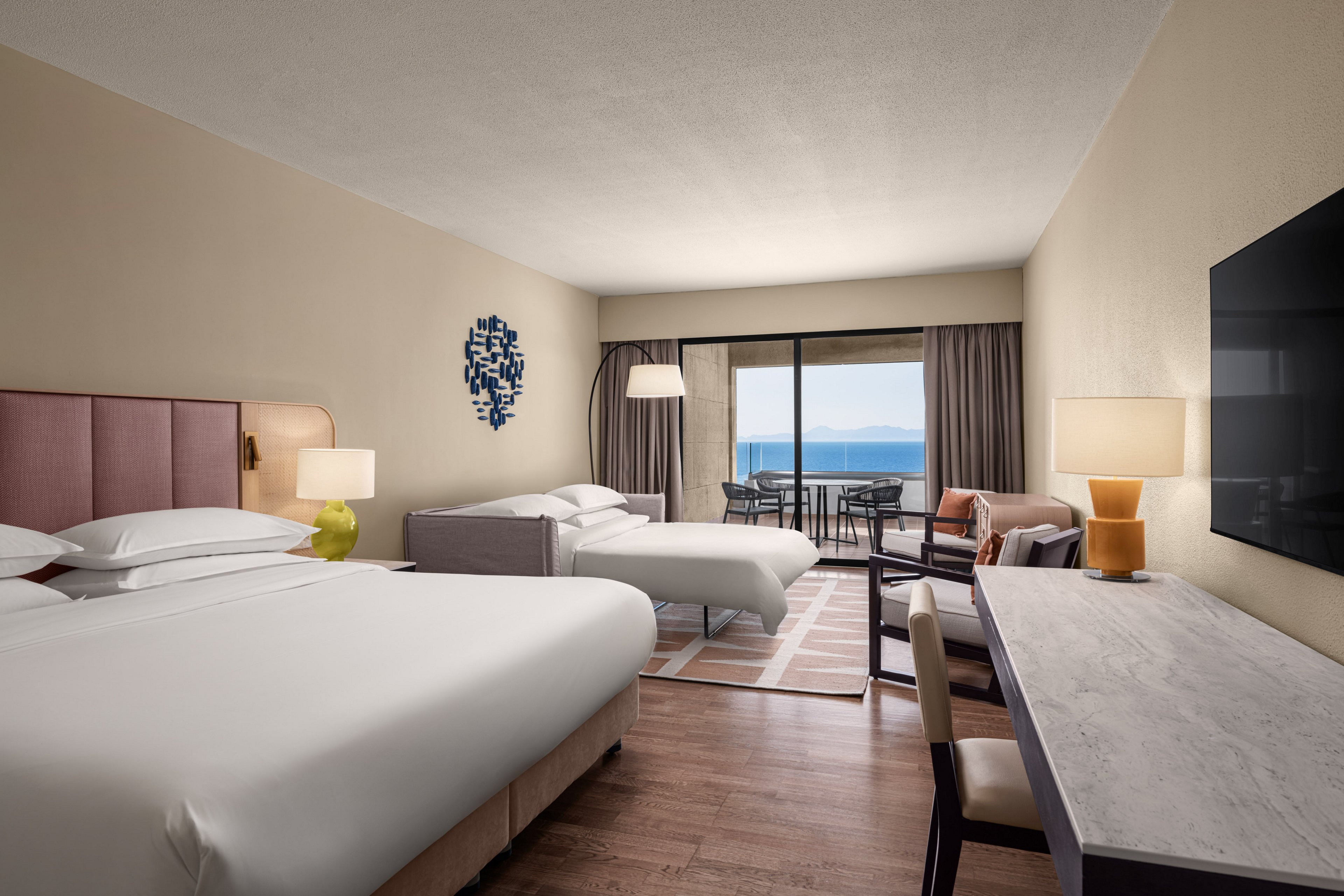 Contemporary hotel room features two large beds and a sleek workspace with a marble-style desk. A seating area with armchairs and a floor lamp leads to a balcony overlooking a calm sea and distant mountains. Neutral walls, wooden flooring, and minimalist decor create a clean, relaxing atmosphere. Large windows and glass doors allow natural light to brighten the interior.