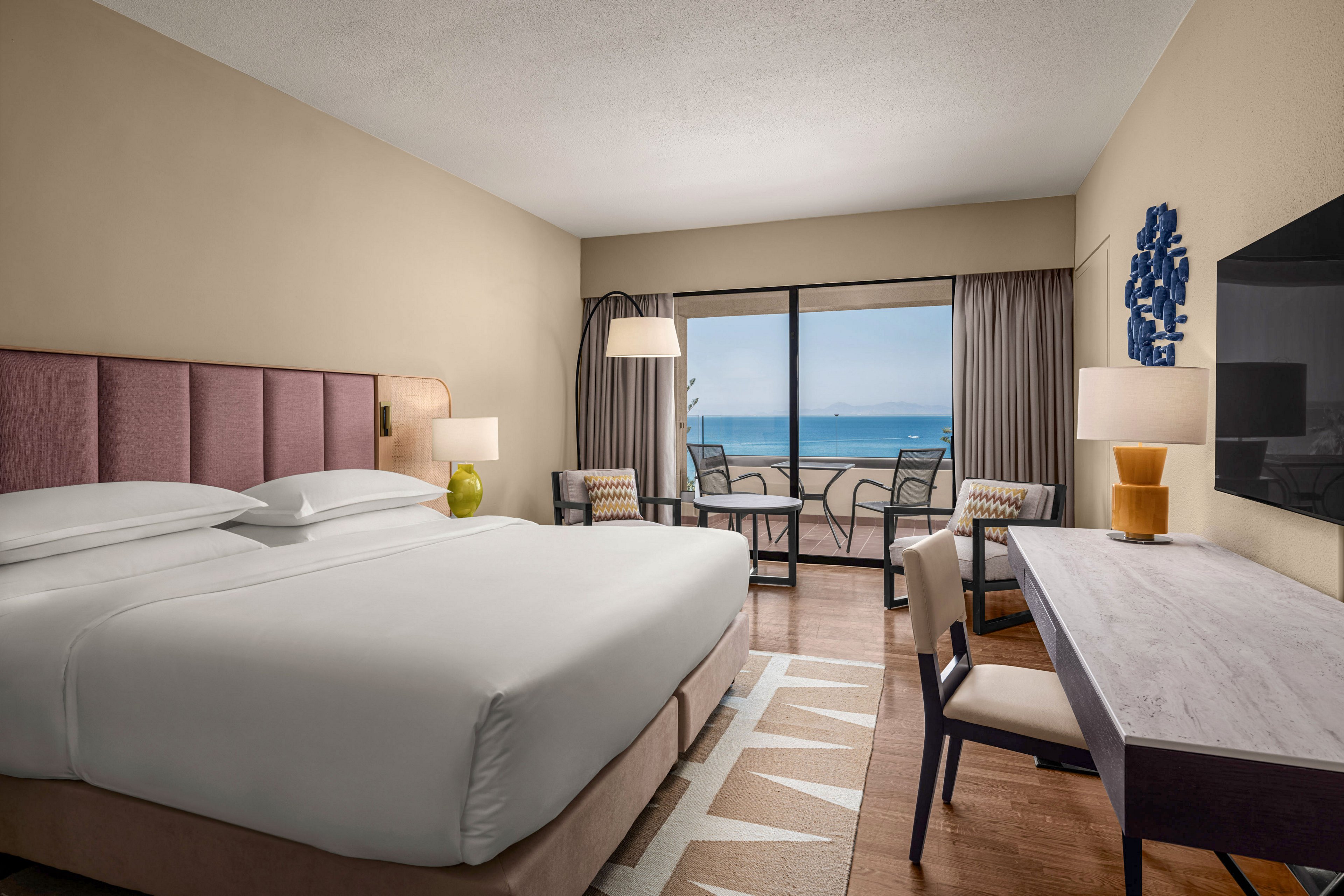 A contemporary hotel bedroom features a large neatly made bed with white linens and a padded headboard. The room includes a desk, chair, and seating area with floor lamp, all facing sliding glass doors that open to a balcony. Outside, a small table and chairs overlook a calm blue ocean and clear sky. Neutral walls, soft lighting, and minimalist decor create a relaxed, inviting atmosphere.