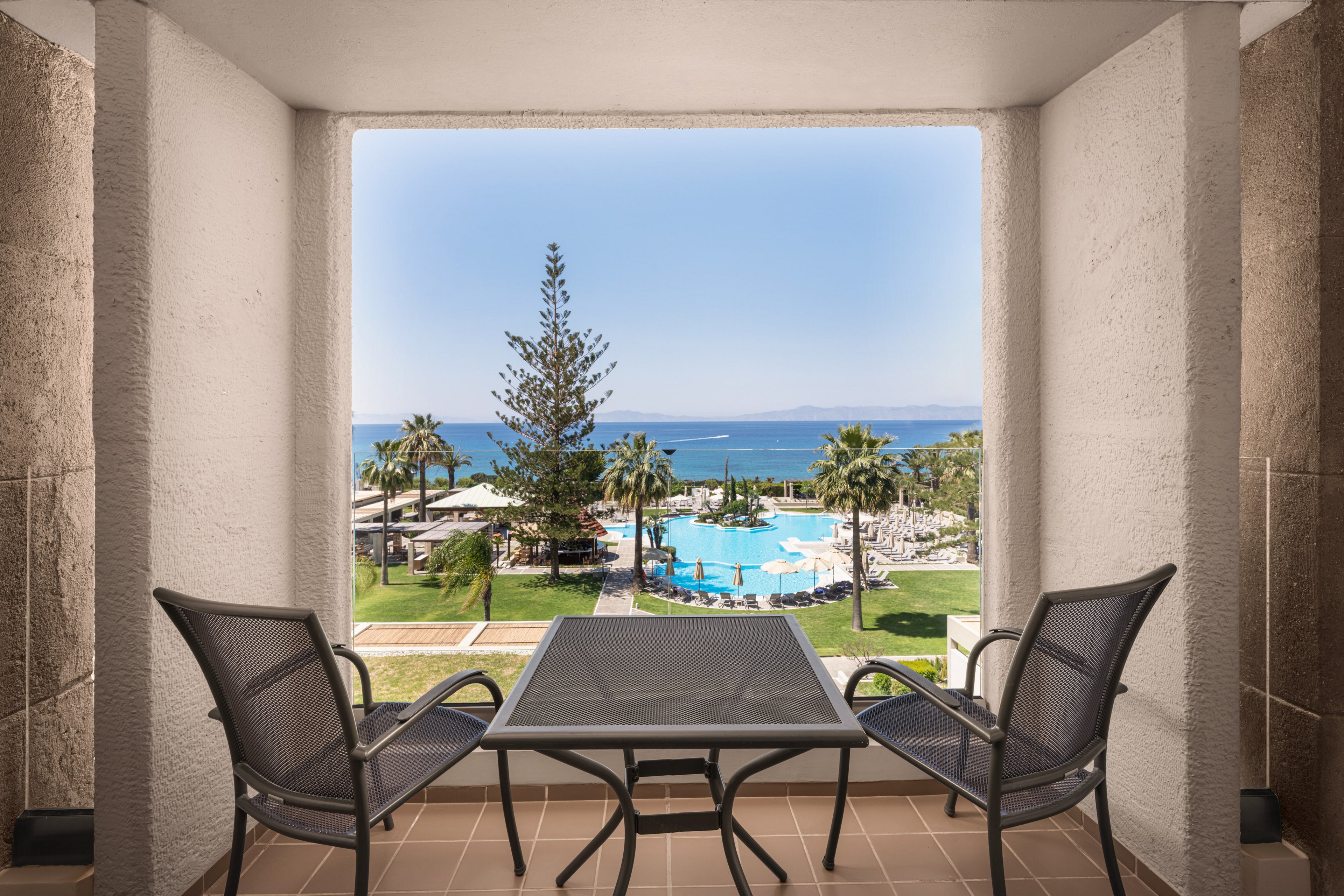 A small balcony with a glass-top table and two mesh chairs overlooks a tropical resort. Beyond the terrace, palm trees surround a bright blue swimming pool with sun loungers and umbrellas. The scene opens out to a calm sea and distant coastline under a clear sky. Warm tones of the balcony contrast with the cool blues of the water, creating a relaxed vacation atmosphere.